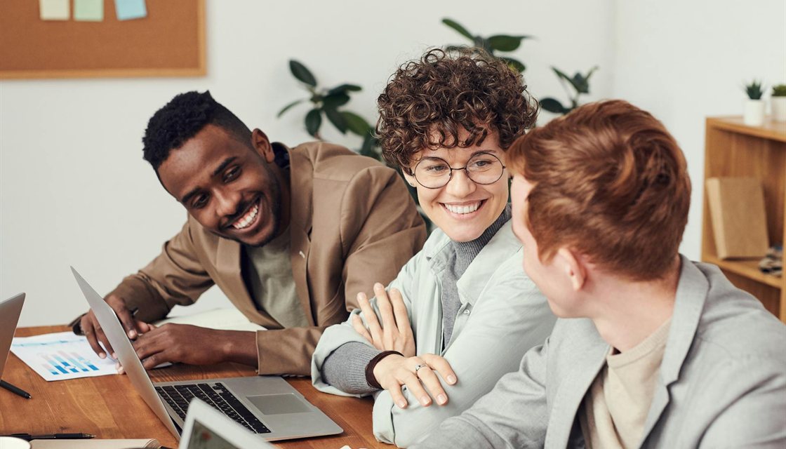 Three colleagues smiling around a laptop and printed charts at a wooden desk