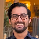 Man with glasses, beard, and mustache smiling indoors with blurred background.