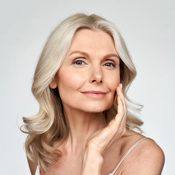 Smiling mature woman with gray hair touching her cheek against a light background.
