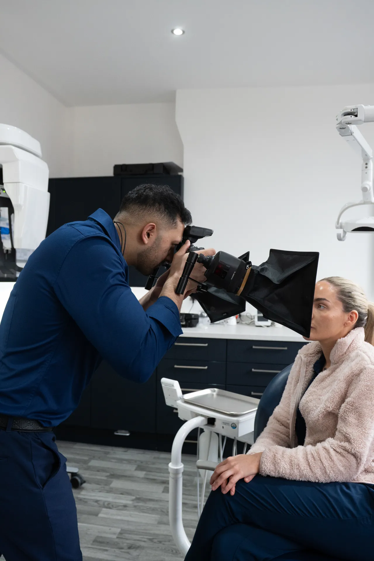 Man in blue shirt using specialized camera equipment to photograph a woman sitting in a dental clinic.