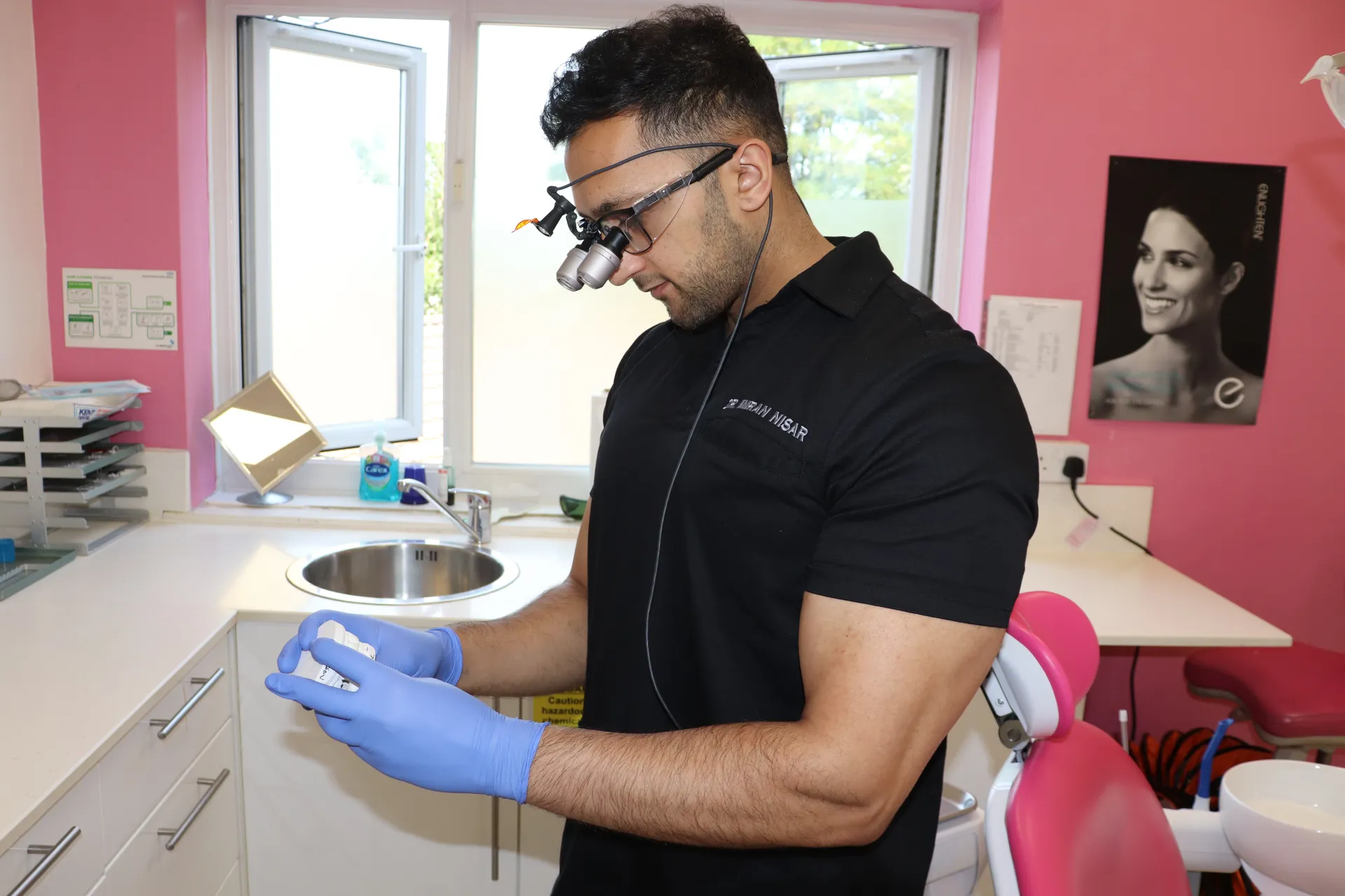A dentist wearing magnifying glasses and blue gloves is examining a dental mold in a clinic with pink walls.