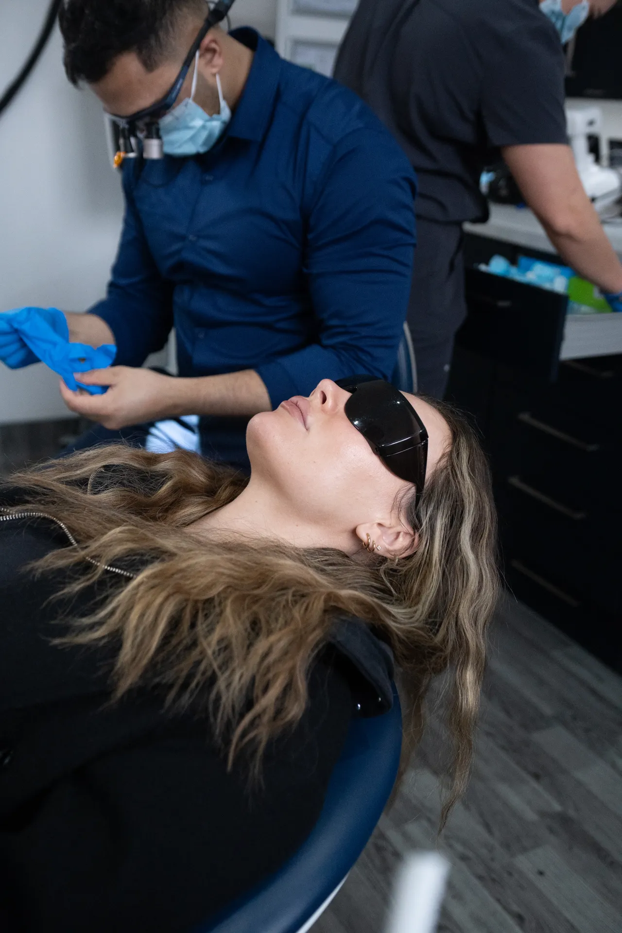 Woman reclining in a dental chair wearing protective dark glasses while a dentist in blue scrubs and face mask prepares with blue gloves.