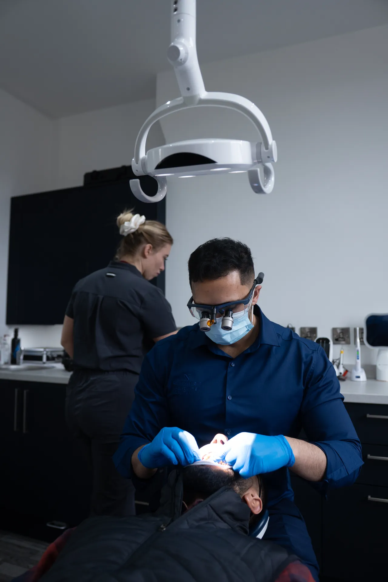 Dentist wearing magnifying glasses and gloves performing a dental procedure on a patient in a dental clinic.
