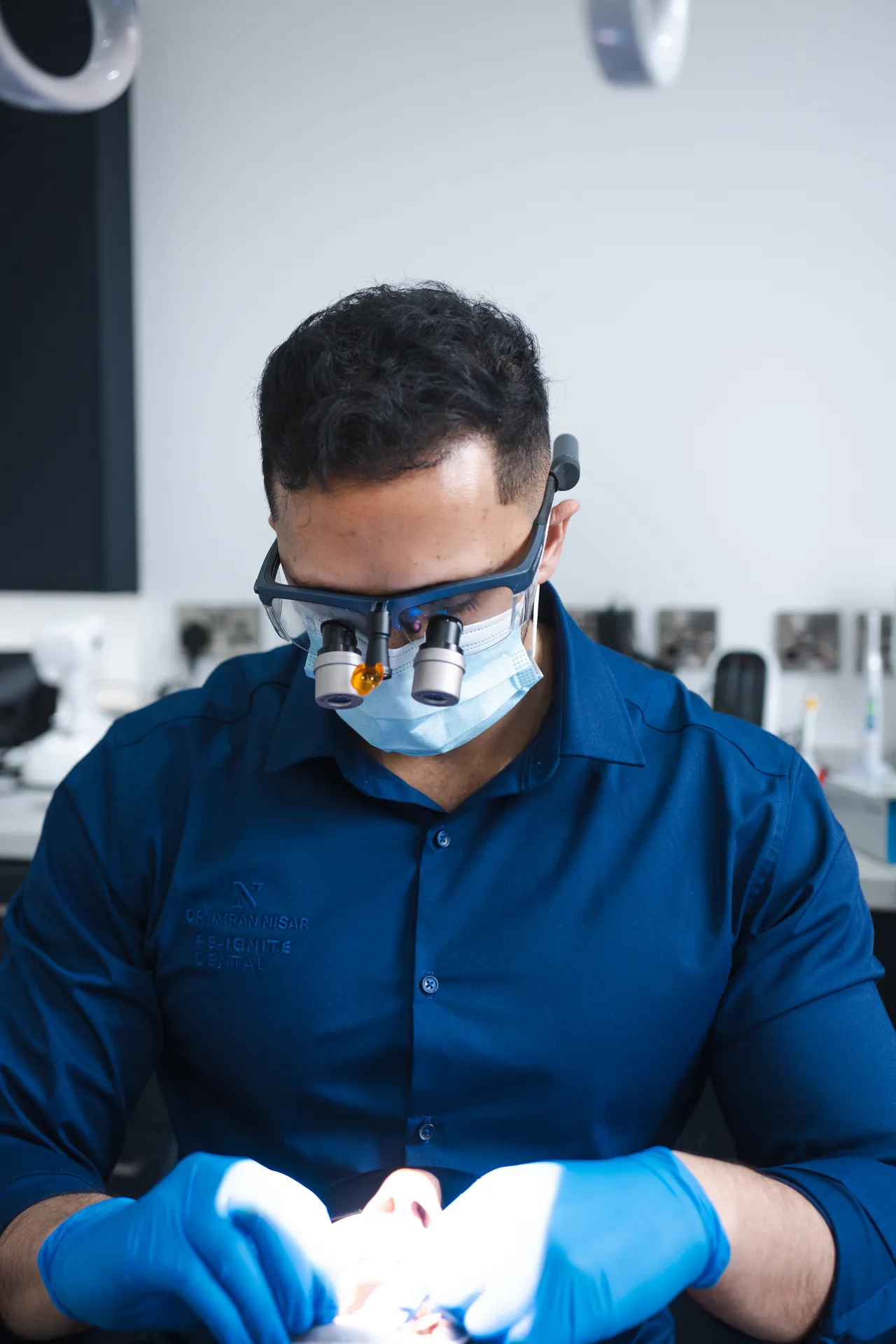 Dentist wearing magnifying loupes, face mask, and blue gloves working on a patient in a dental clinic.