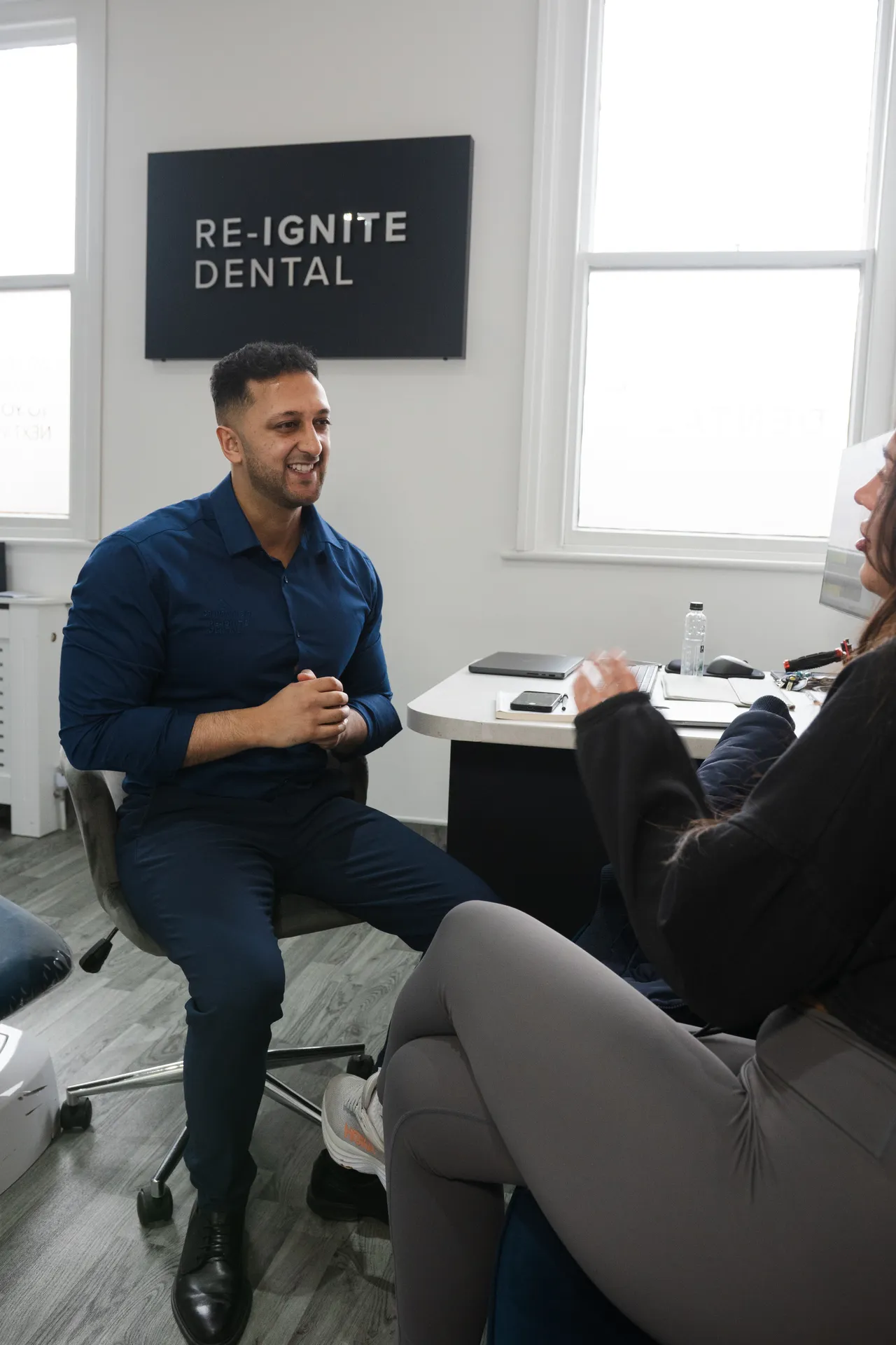 Dentist in a blue shirt smiling and talking with a female patient in a dental office with a Re-Ignite Dental sign on the wall.
