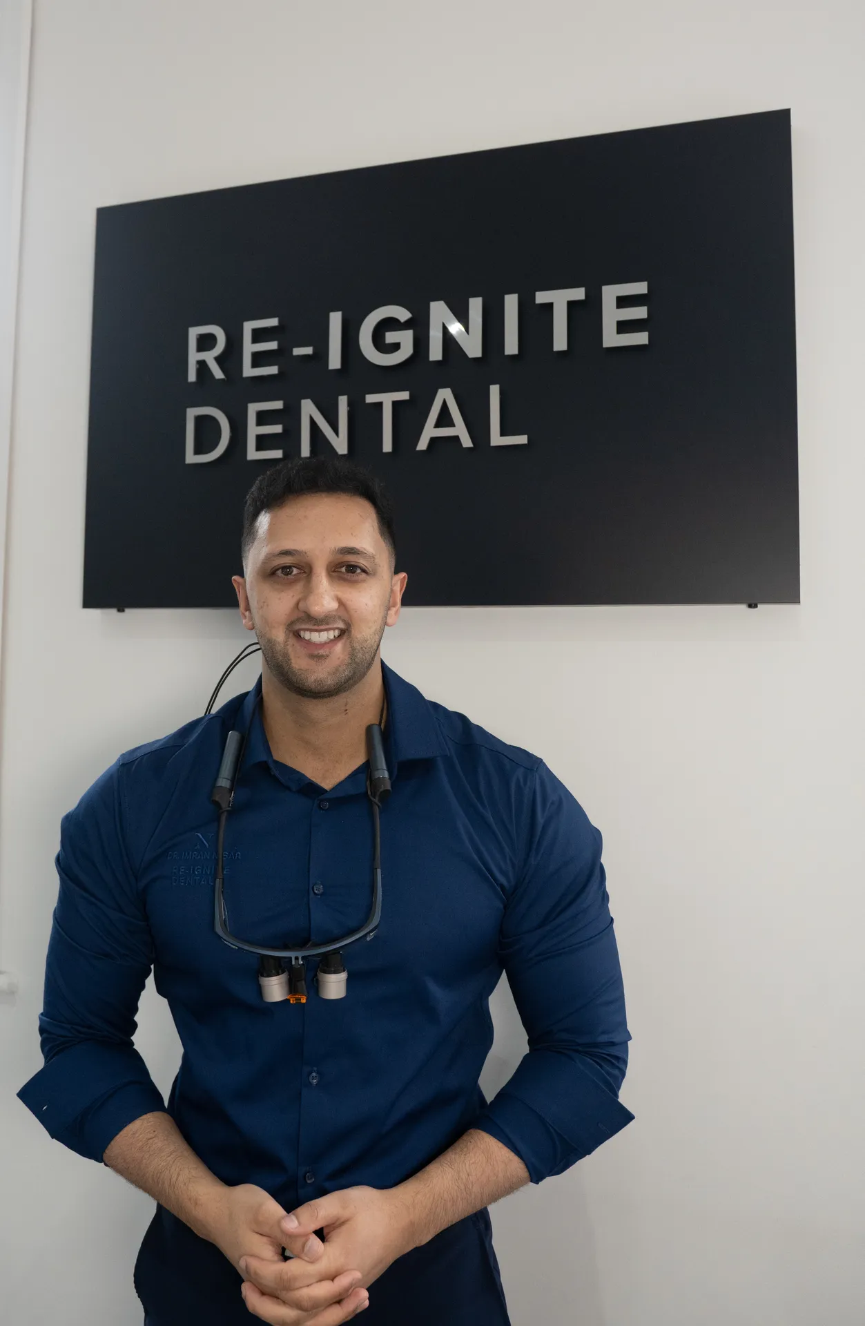 Smiling male dentist in a navy blue shirt standing in front of a black sign that reads 'RE-IGNITE DENTAL.'