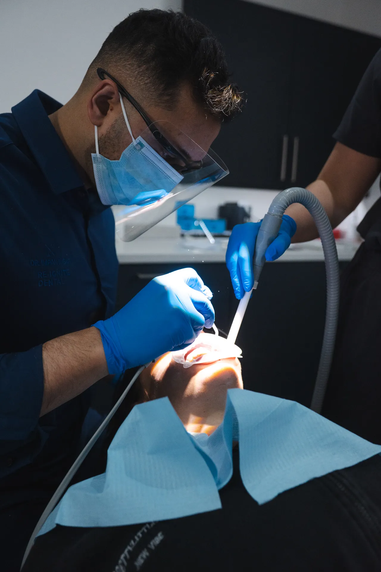 Dentist wearing protective face shield and mask performing dental procedure on patient with blue gloves and medical tools.