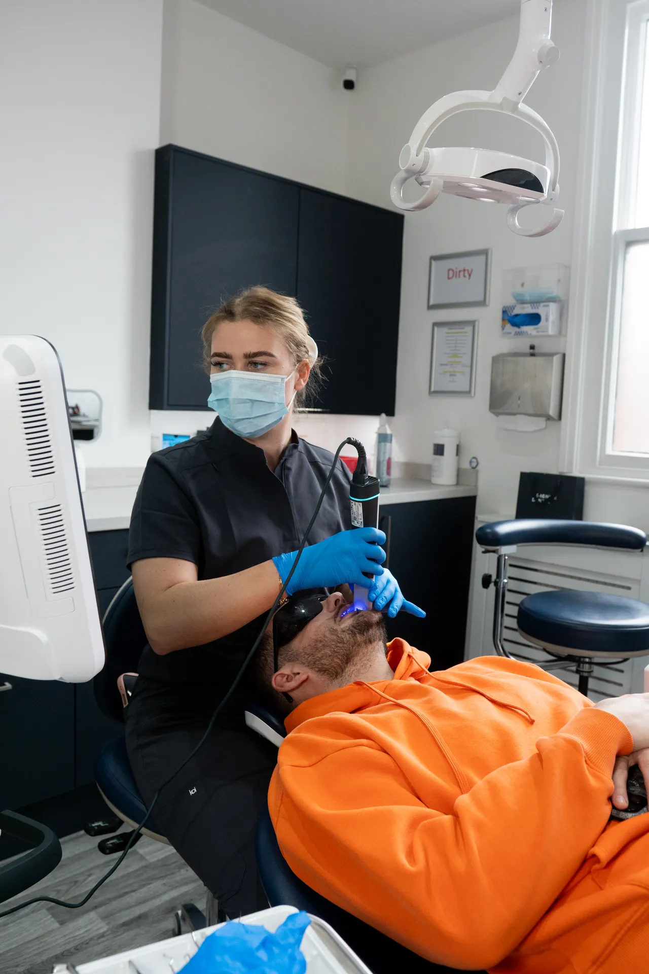 Dentist wearing a mask and gloves using a dental tool with blue light on a patient in an orange hoodie lying in a dental chair.