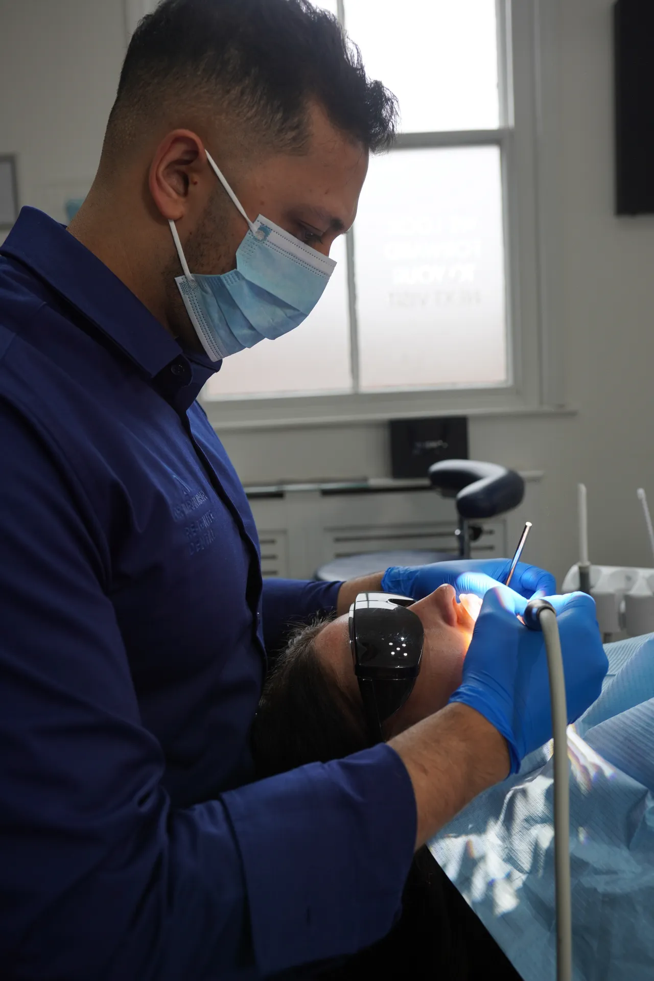Dentist wearing a surgical mask and gloves examining a patient's mouth with dental tools in a clinic.