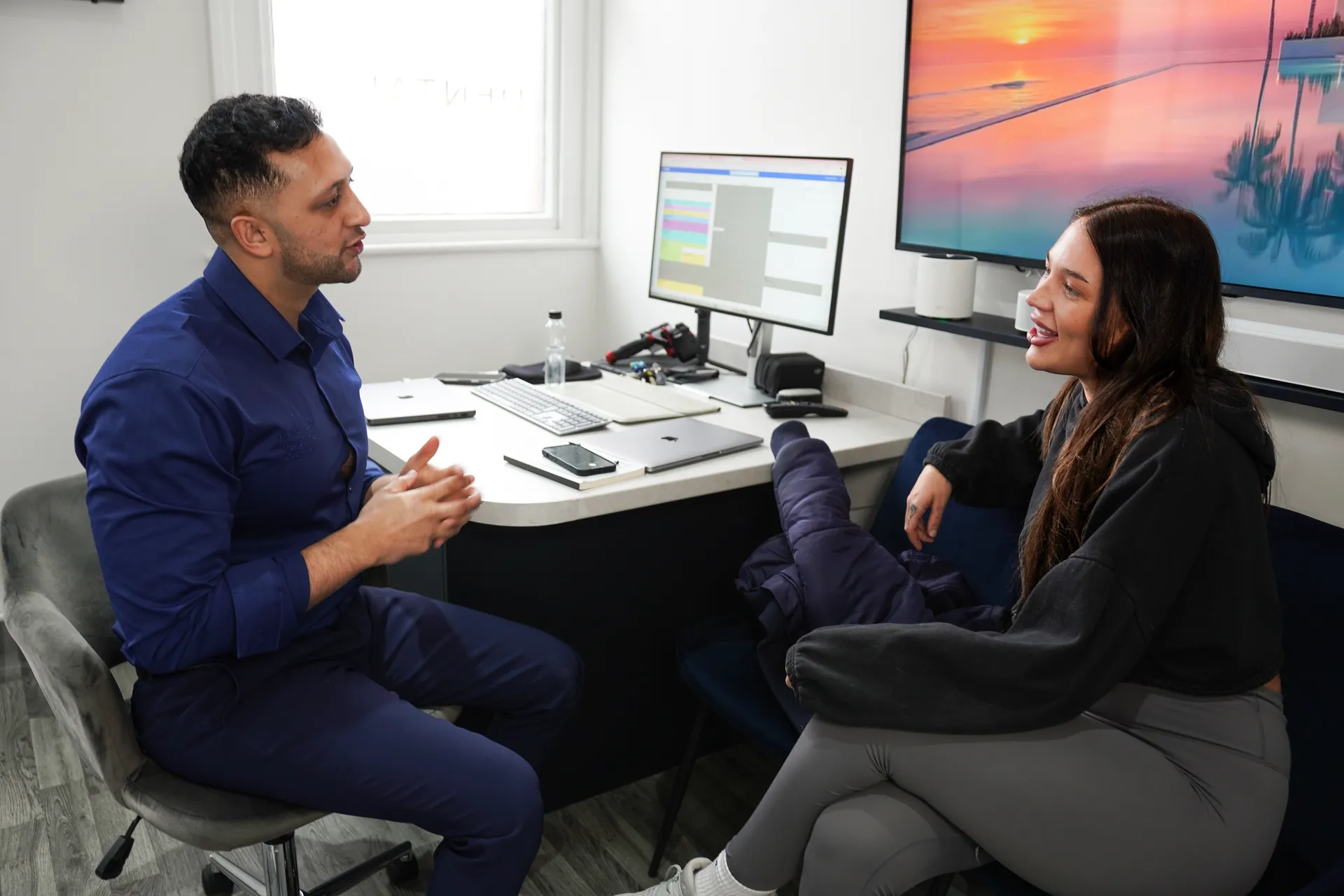 Man in blue shirt and woman in black hoodie sitting and talking face-to-face in an office with computer screens.
