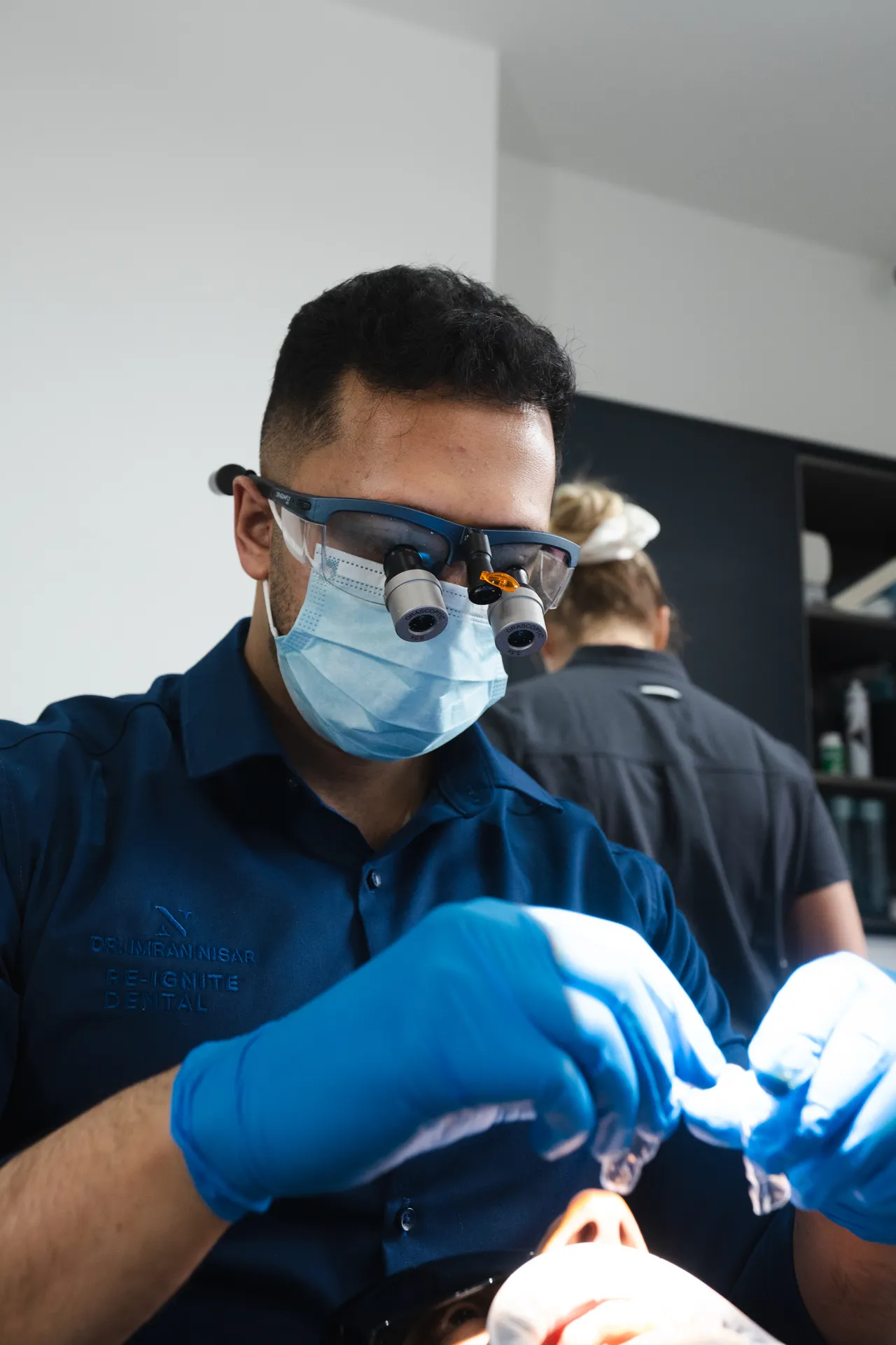 Dentist wearing magnifying glasses and blue gloves working on a patient's mouth in a clinical setting.