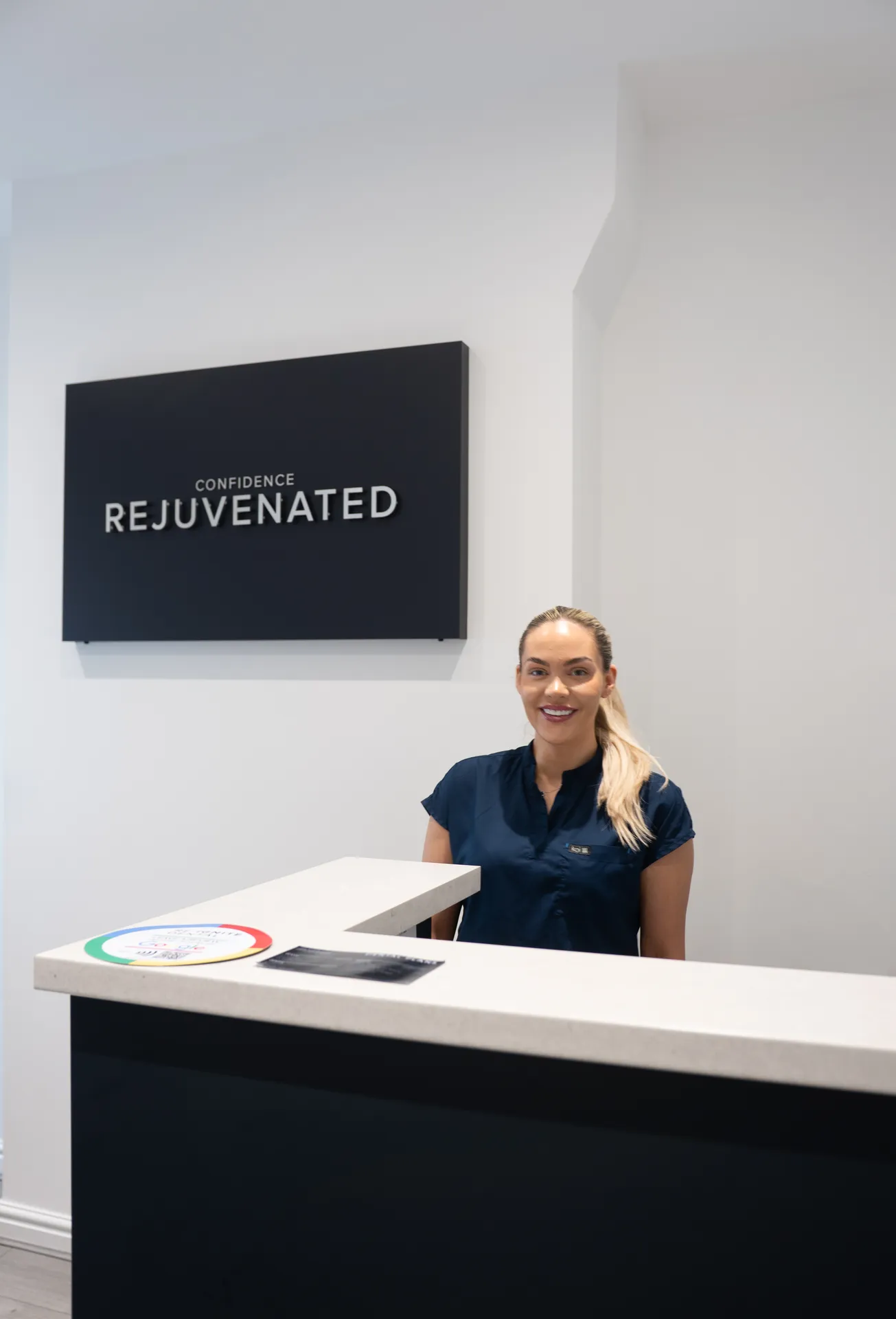 Smiling woman in navy scrubs standing behind a white reception desk with a black sign on the wall reading 'Confidence Rejuvenated'.