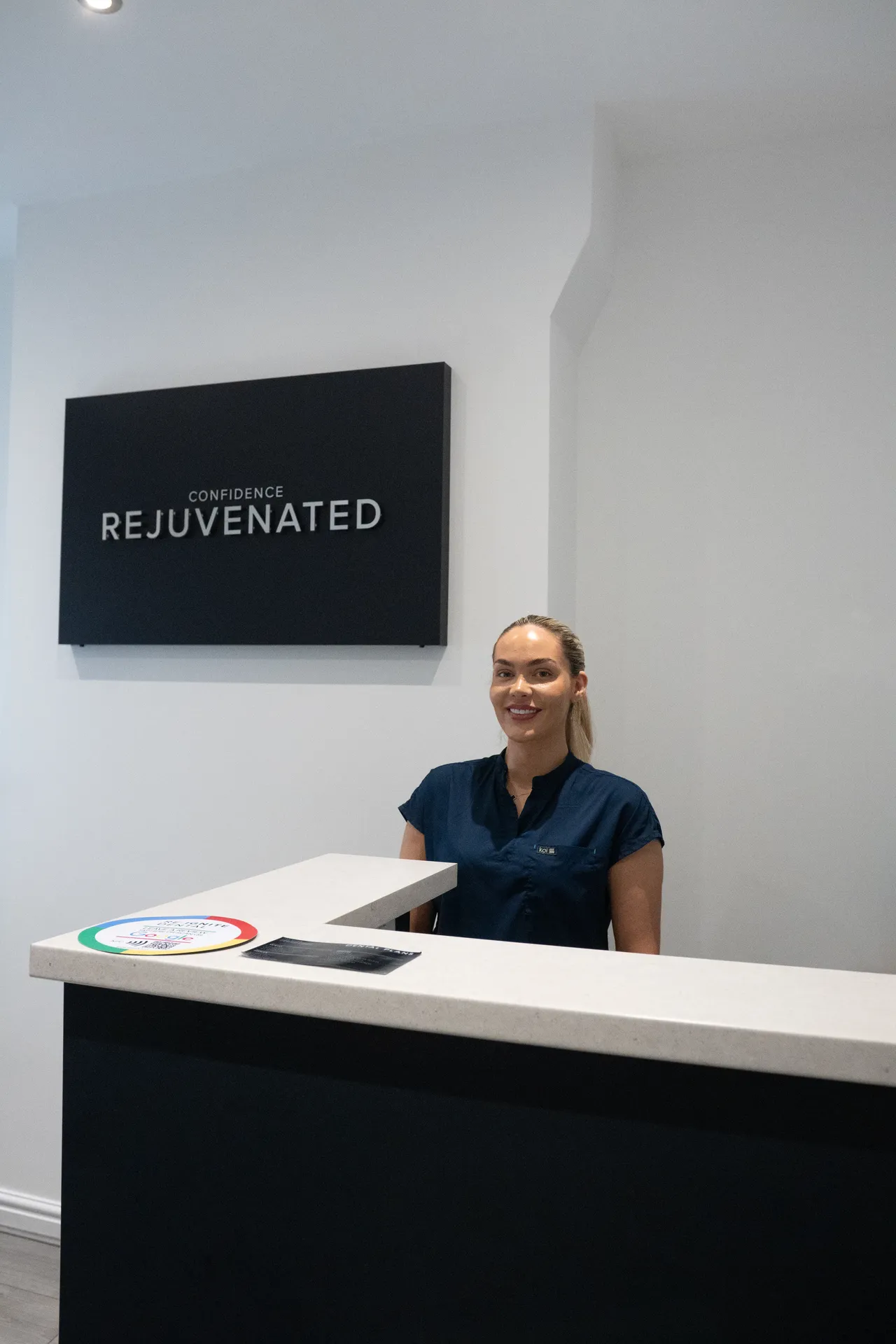 Receptionist smiling behind a modern white and black reception desk with a sign that reads 'Confidence Rejuvenated' on the wall behind.