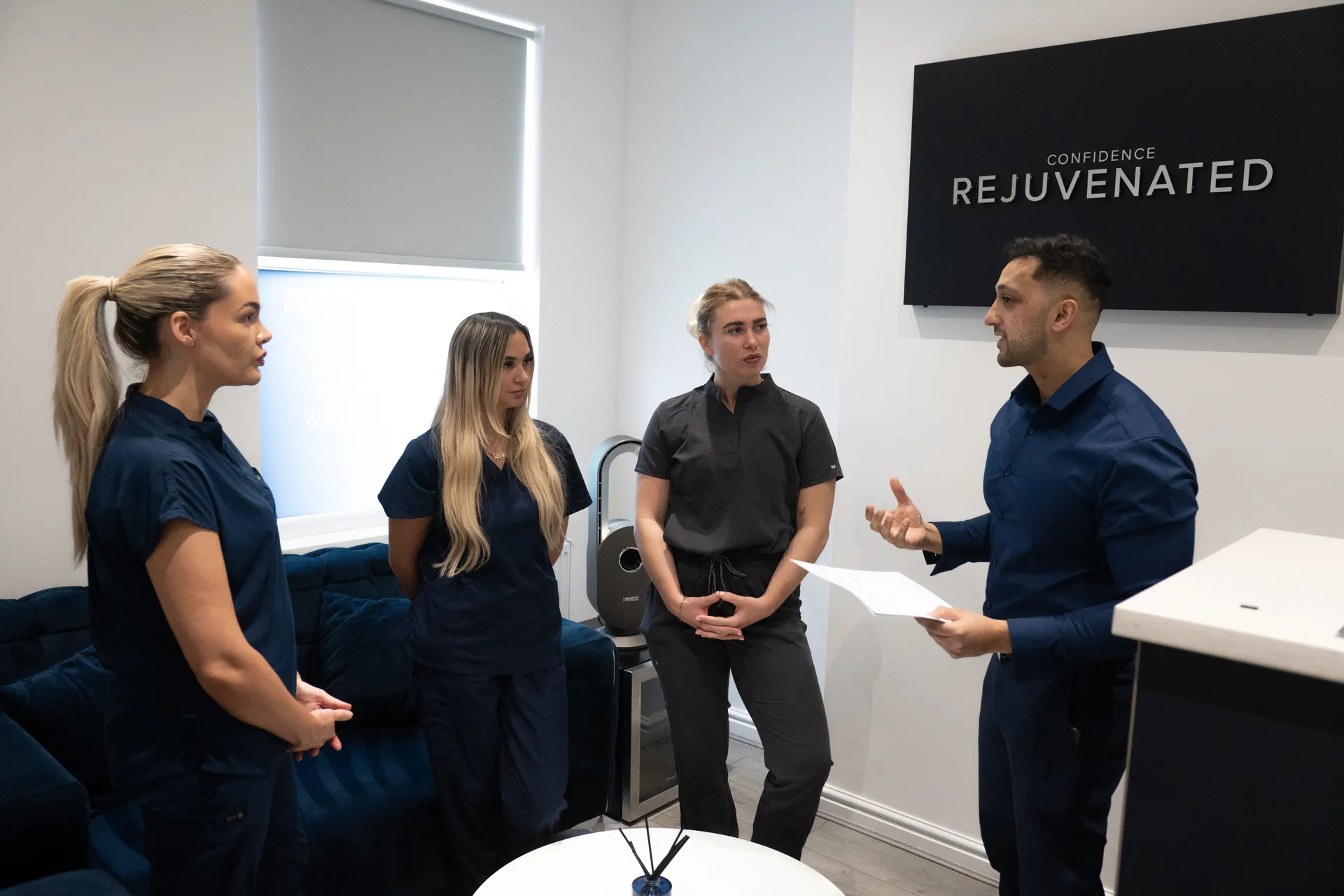 Three women in dark scrubs listening to a man in a blue shirt holding a paper in a modern office with a sign that reads 'Confidence Rejuvenated'.