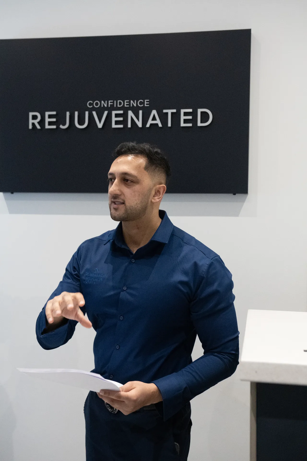 Man in a navy blue shirt holding papers and gesturing, standing in front of a sign that reads 'Confidence Rejuvenated'.