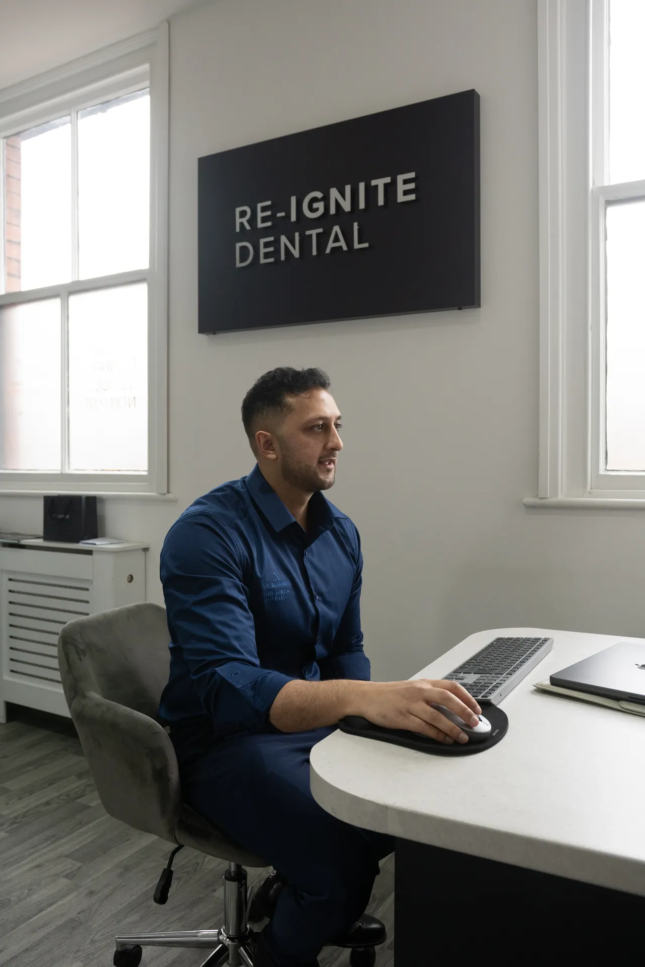 Man in navy blue shirt sitting at a desk using a computer mouse in a dental office with a RE-IGNITE DENTAL sign on the wall.