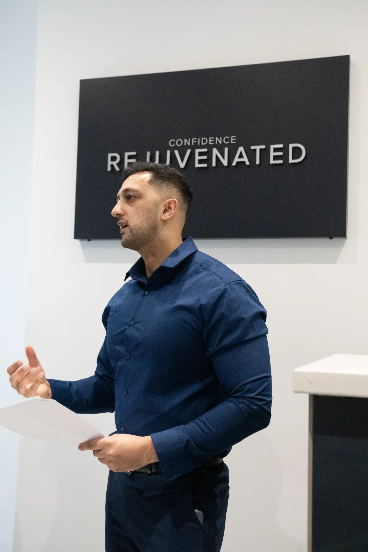 Man in a blue shirt speaking with a paper in his hand in front of a sign that reads 'Confidence Rejuvenated'.
