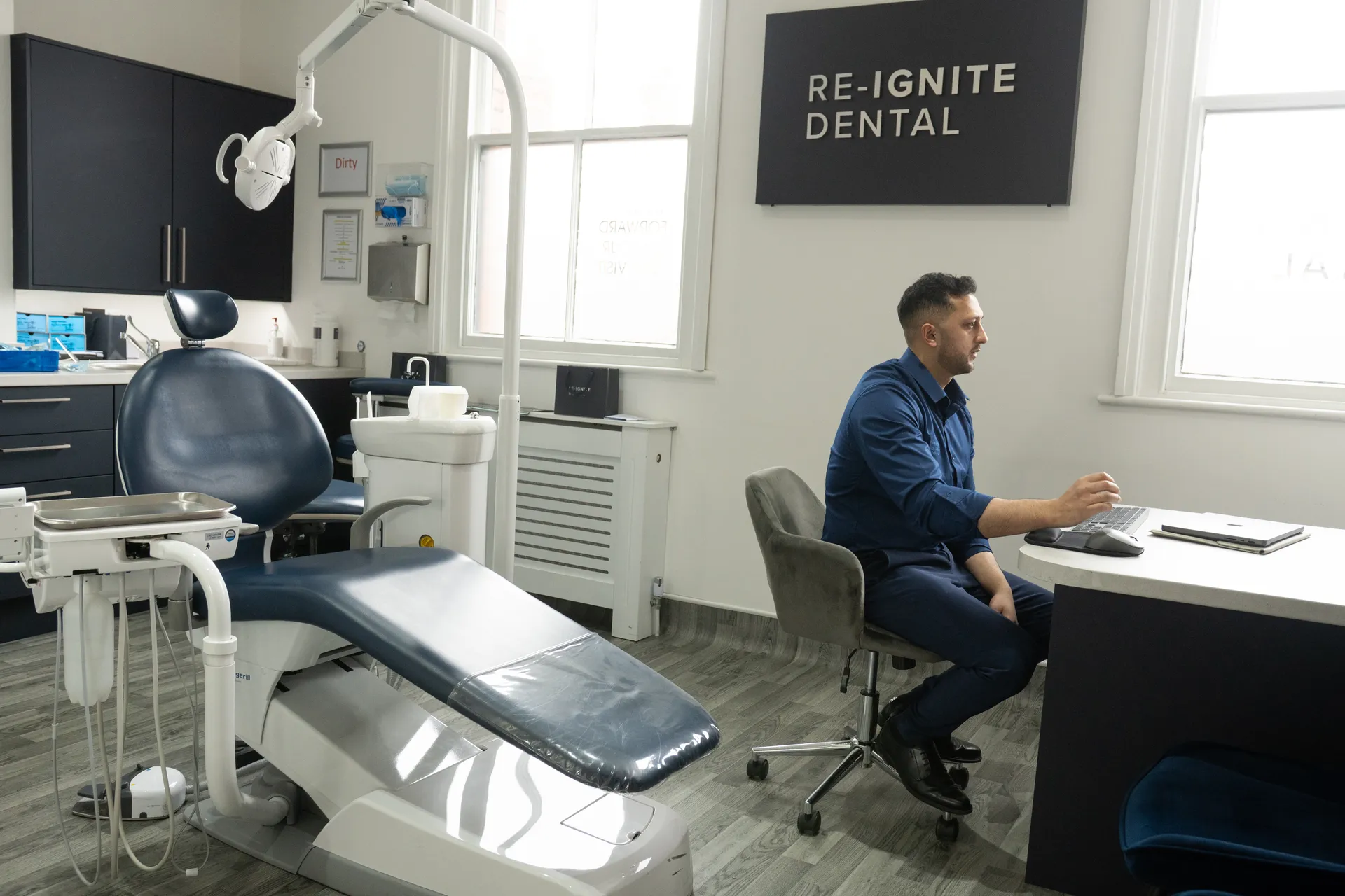 Dentist seated at a desk in a modern dental office with dental chair and equipment nearby.