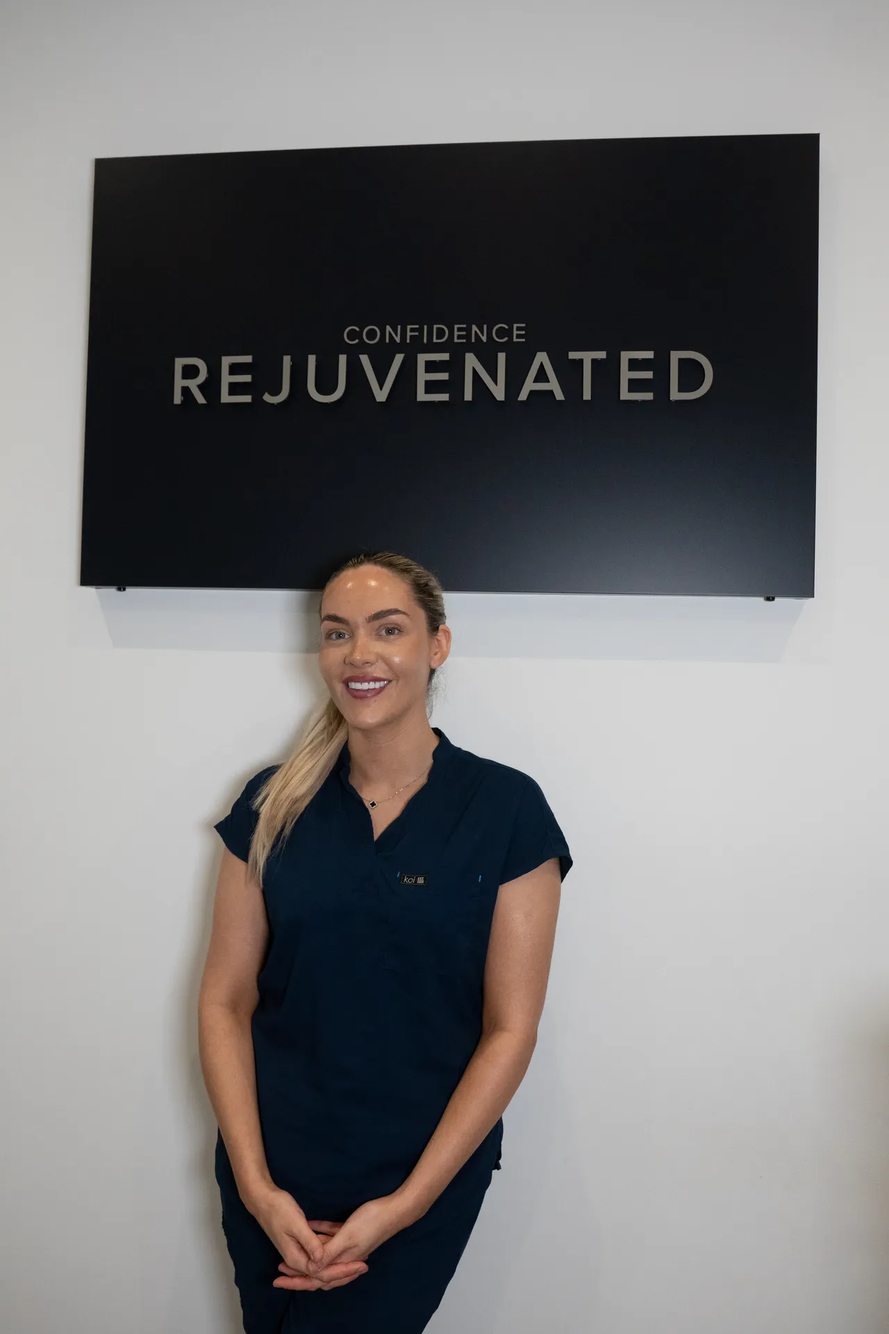 Smiling woman in navy scrubs standing in front of a sign that reads Confidence Rejuvenated.