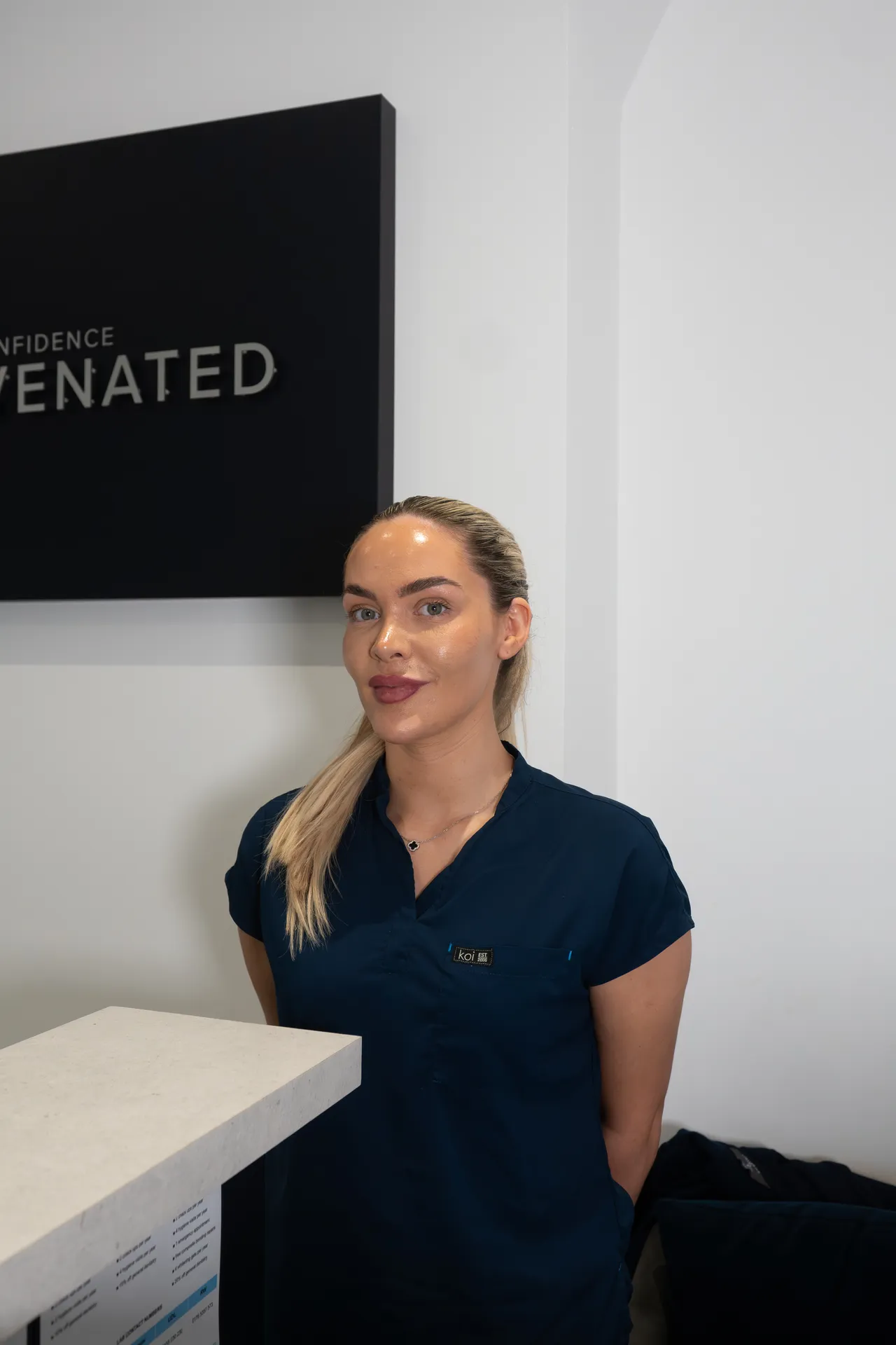 Woman with blonde hair tied back wearing a dark blue scrub top standing behind a counter with a white wall and part of a black sign in the background.