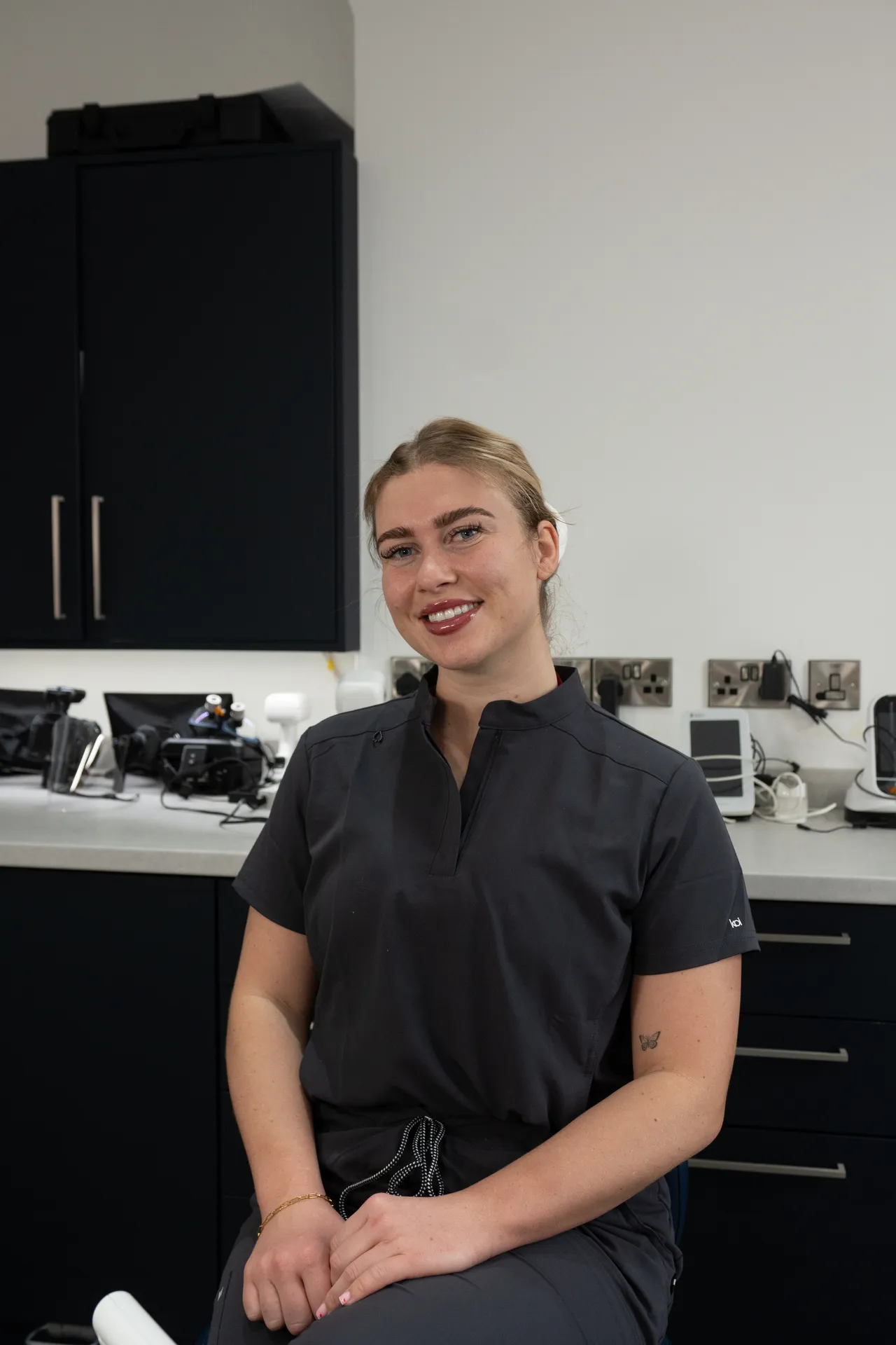 Smiling young woman with blonde hair in dark scrubs sitting in a modern medical or dental office.