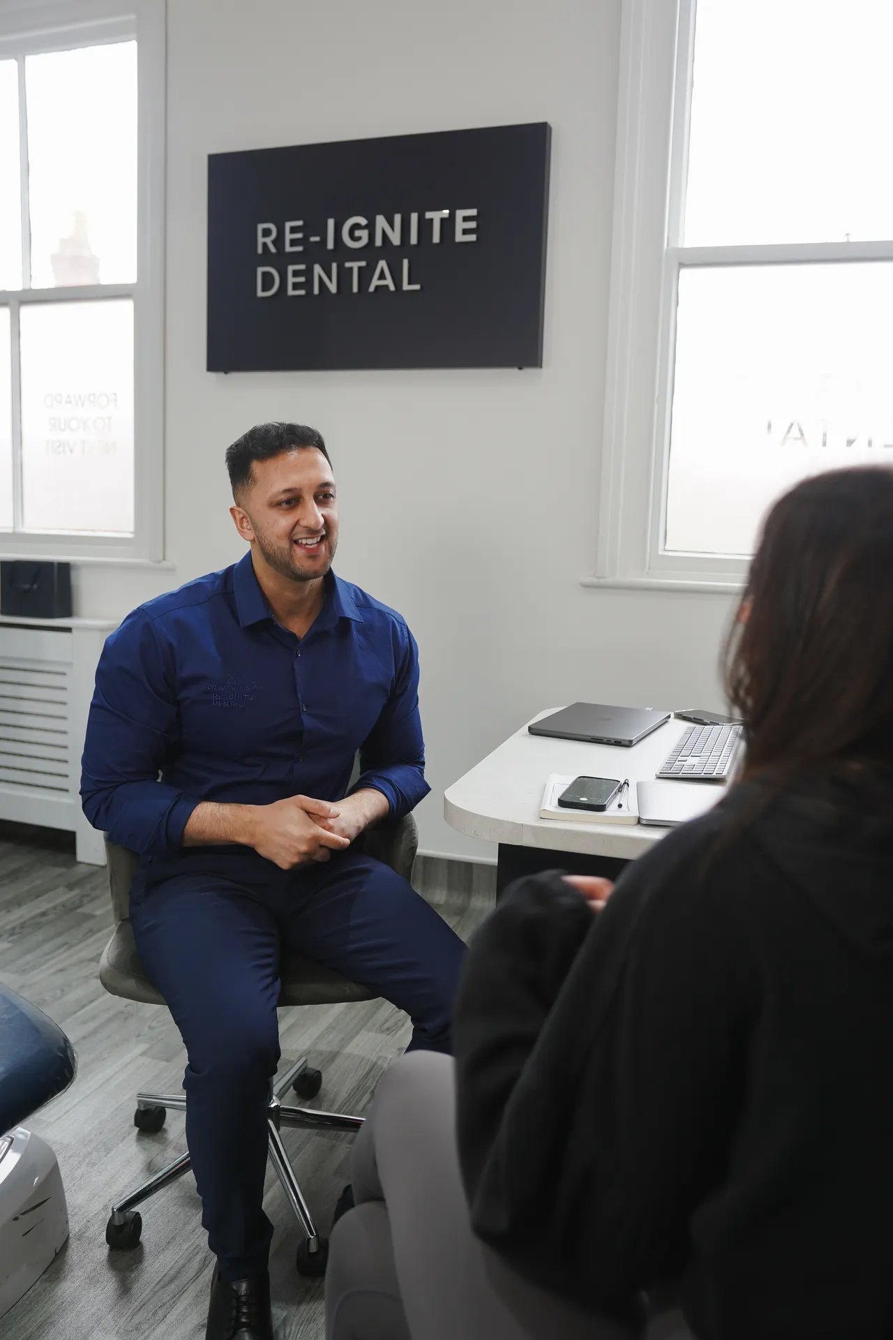 A dentist in navy blue scrubs talking and smiling with a seated patient in a dental office with Re-Ignite Dental sign on the wall.