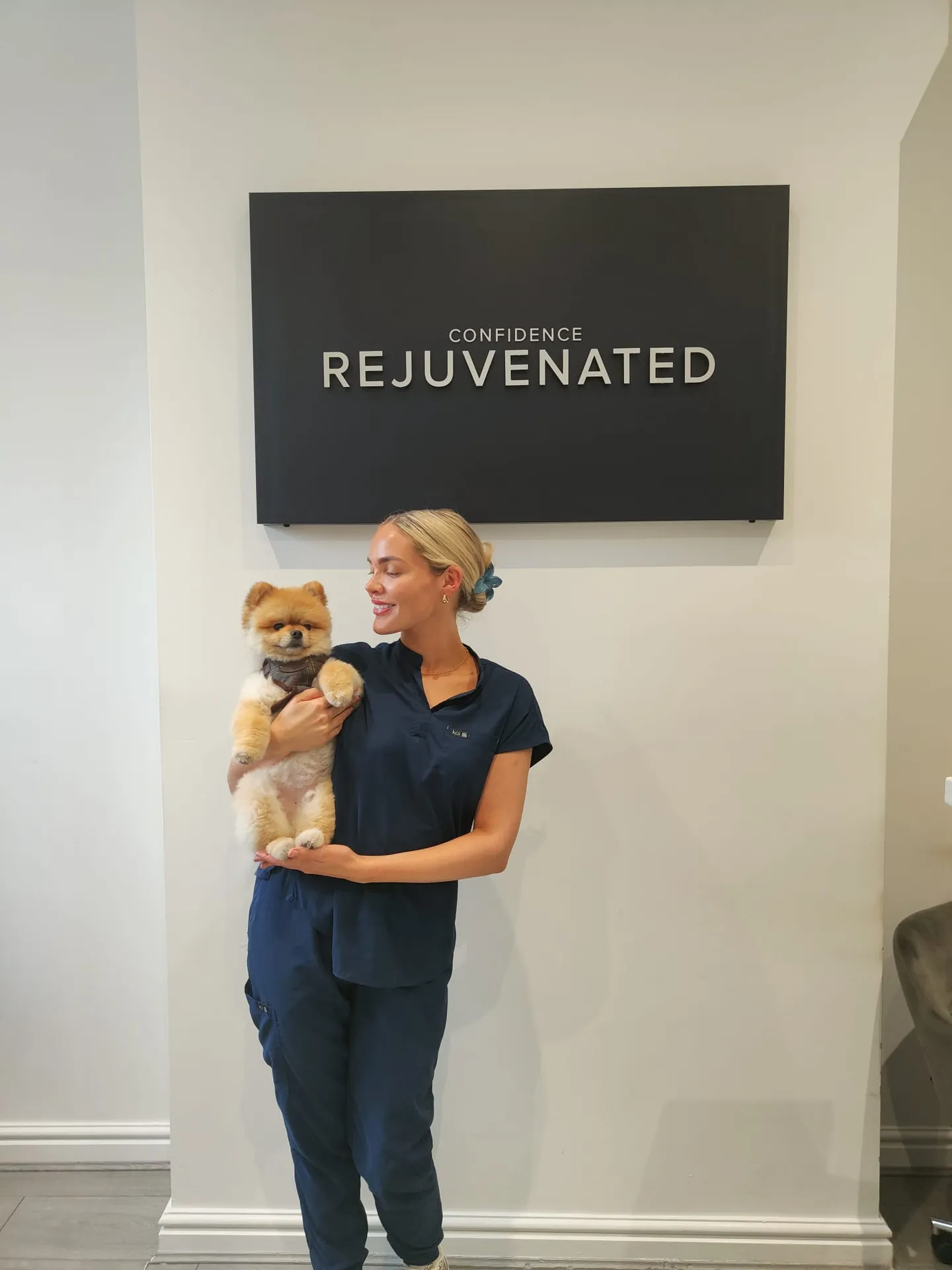 Woman in navy scrubs holding a small fluffy dog under a sign that reads 'Confidence Rejuvenated.'