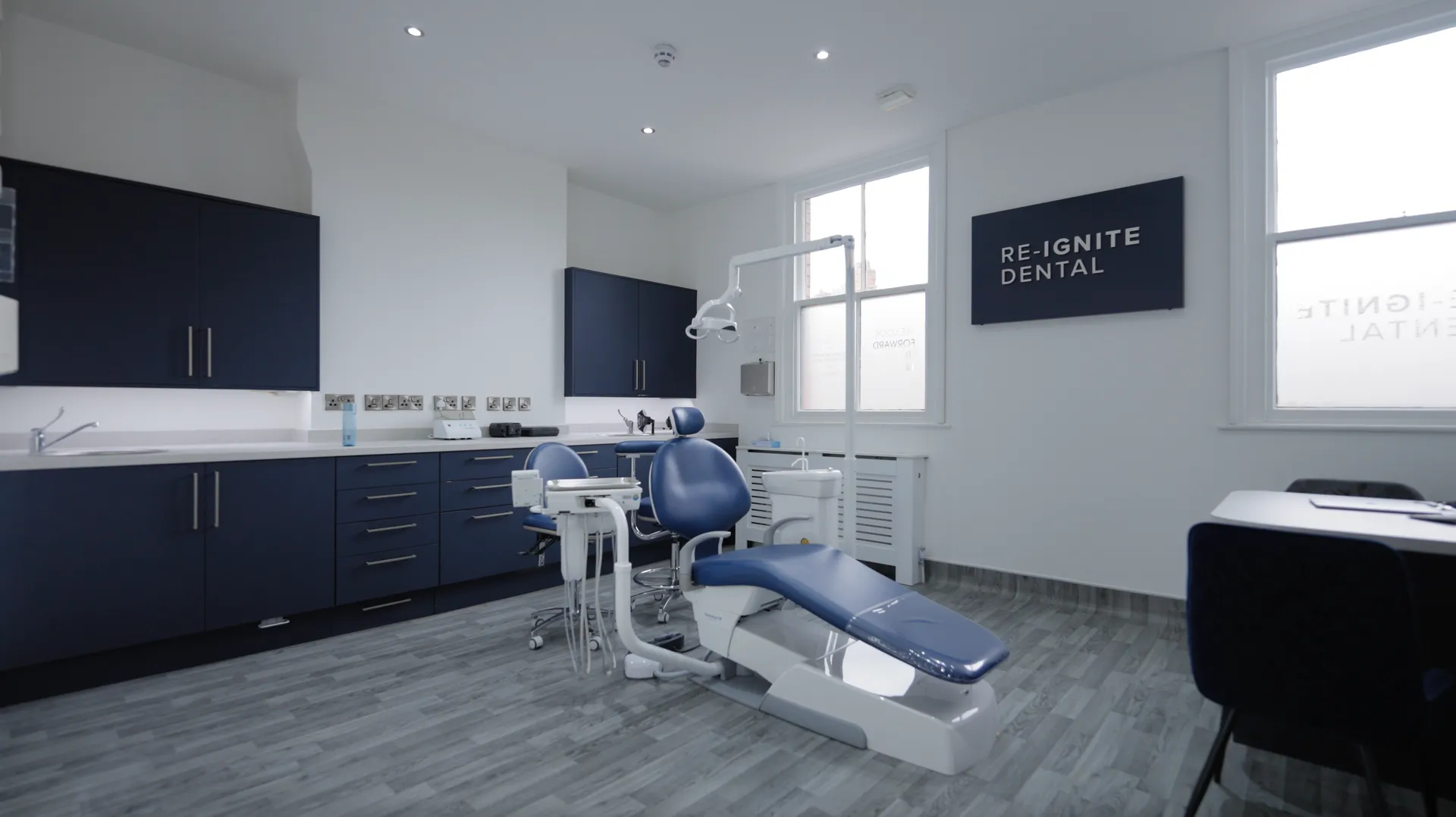 Modern dental treatment room with blue dental chair, cabinetry, and equipment under bright ceiling lights.