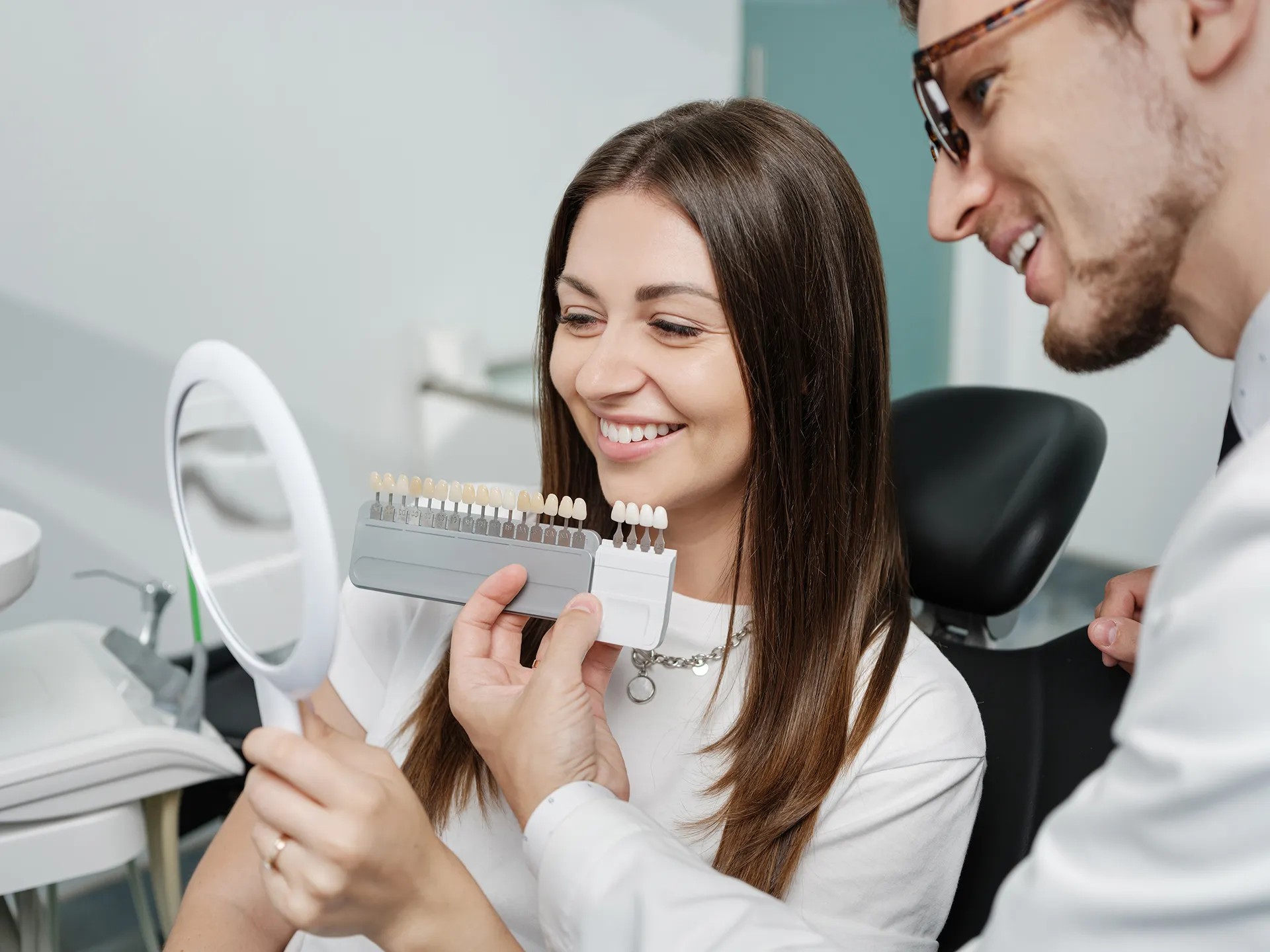 Dentist helping a smiling woman choose veneer shade using a shade guide while she looks in a handheld mirror.