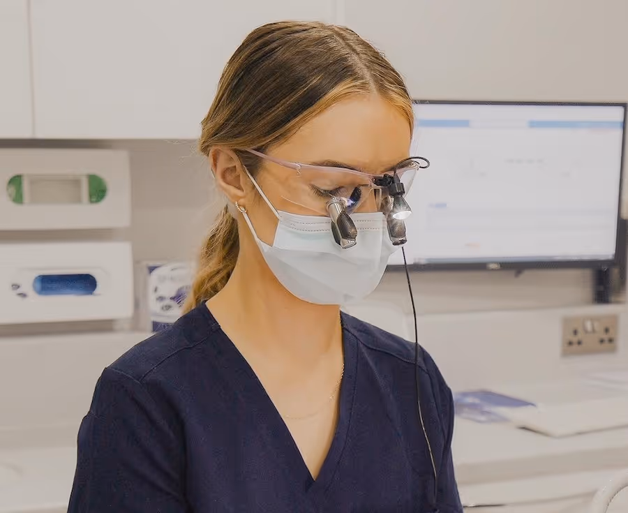 Female medical professional wearing a mask and magnifying glasses with lights in a clinical setting.