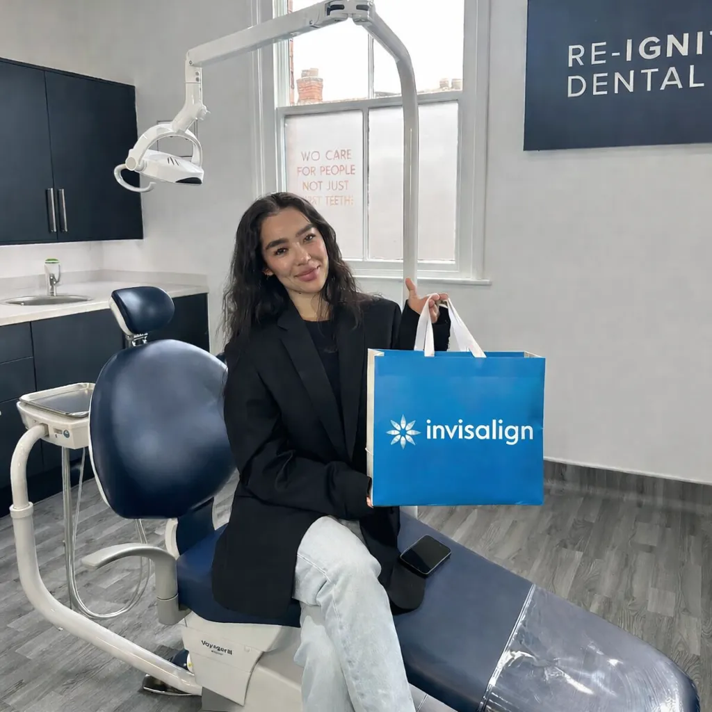 Smiling woman sitting on a dental chair holding a blue Invisalign bag inside a dental office.