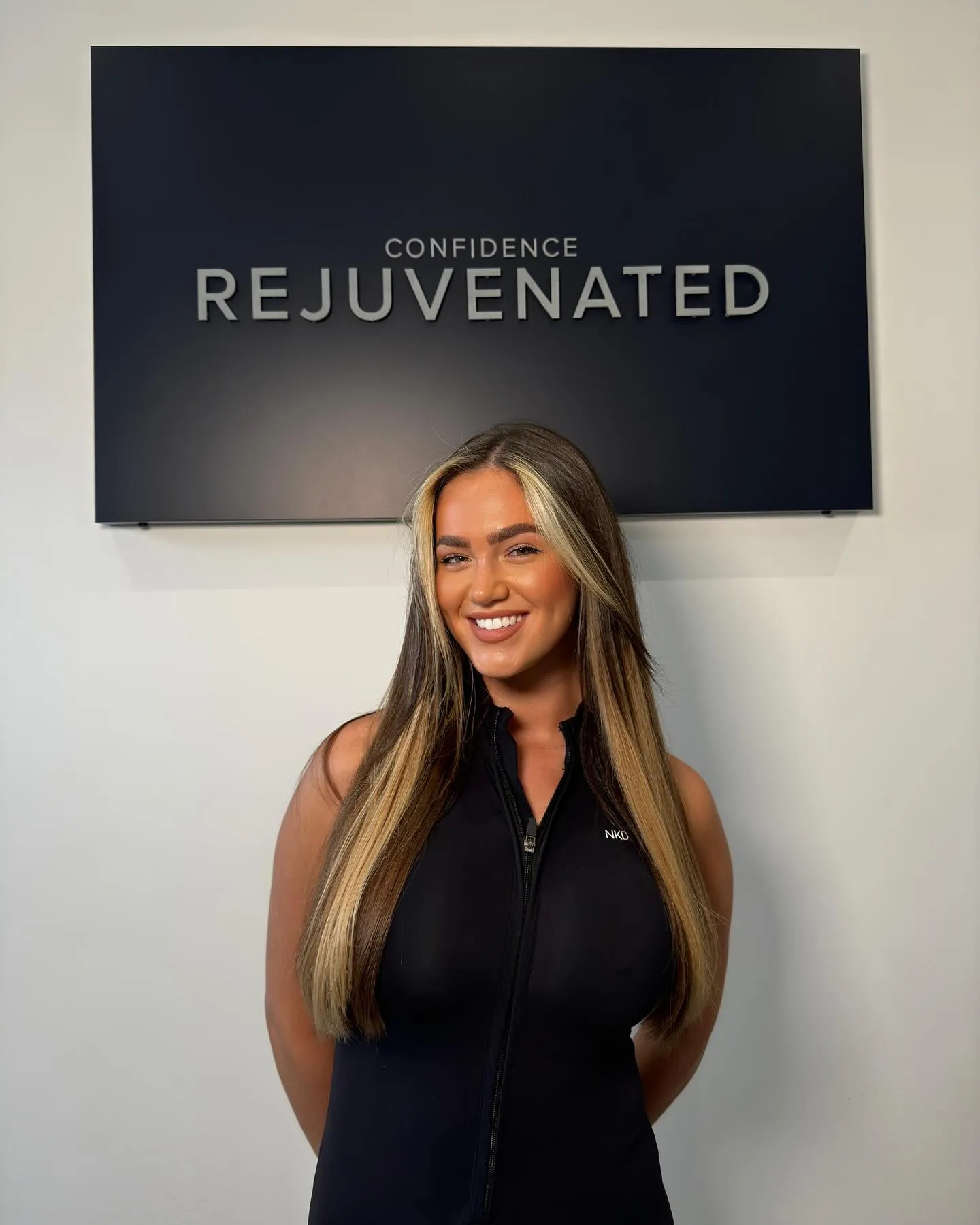 Smiling woman with long blonde highlights stands in front of a sign reading Confidence Rejuvenated.