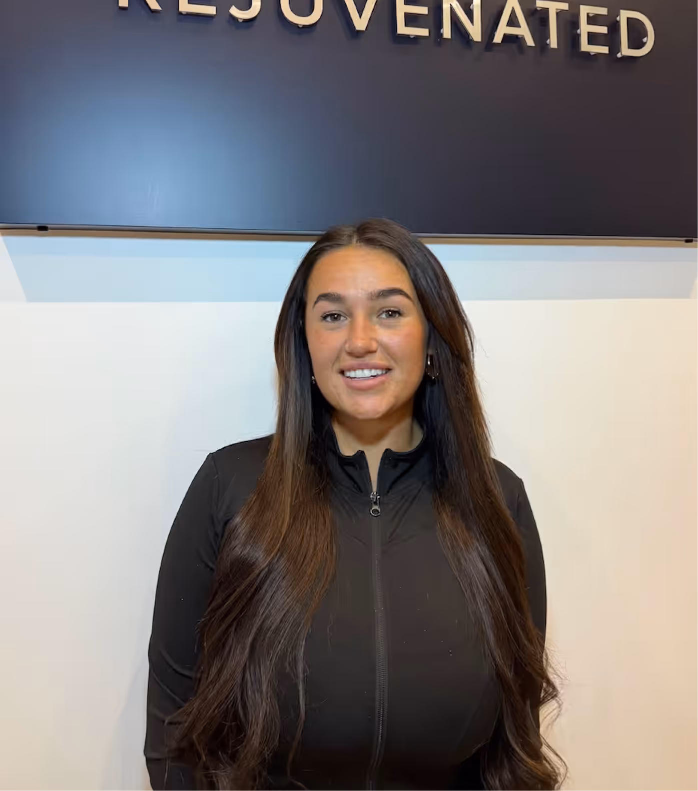 Smiling woman with long dark hair wearing a black zip-up top standing in front of a wall with 'REJUVENATED' sign.