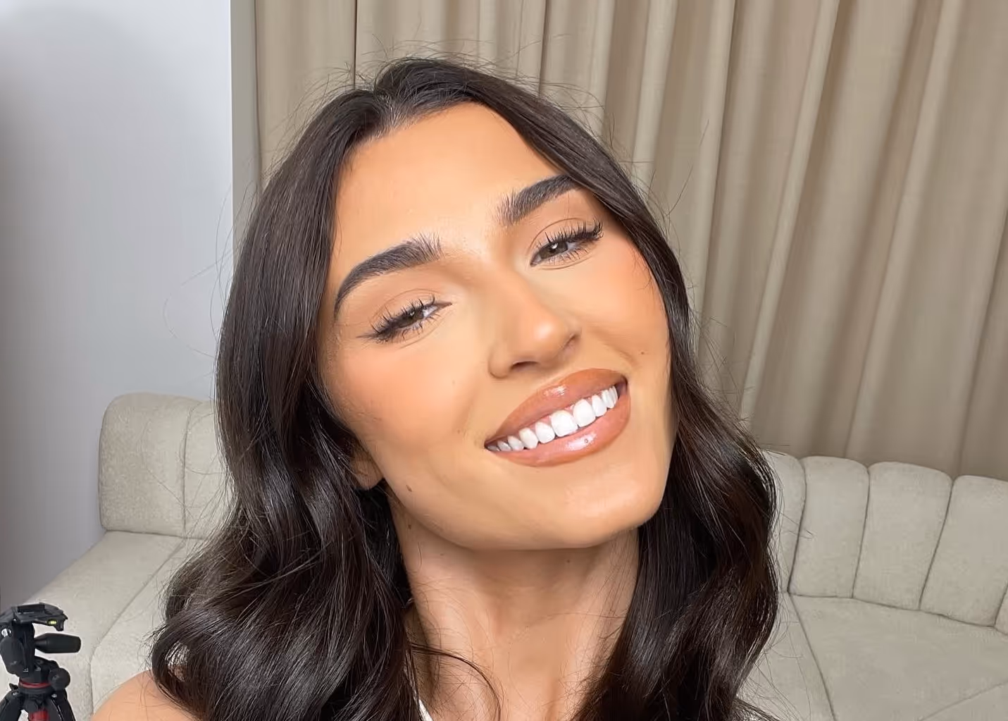 Smiling woman with long dark wavy hair wearing a white top, posing indoors with beige curtains and sofa in background.