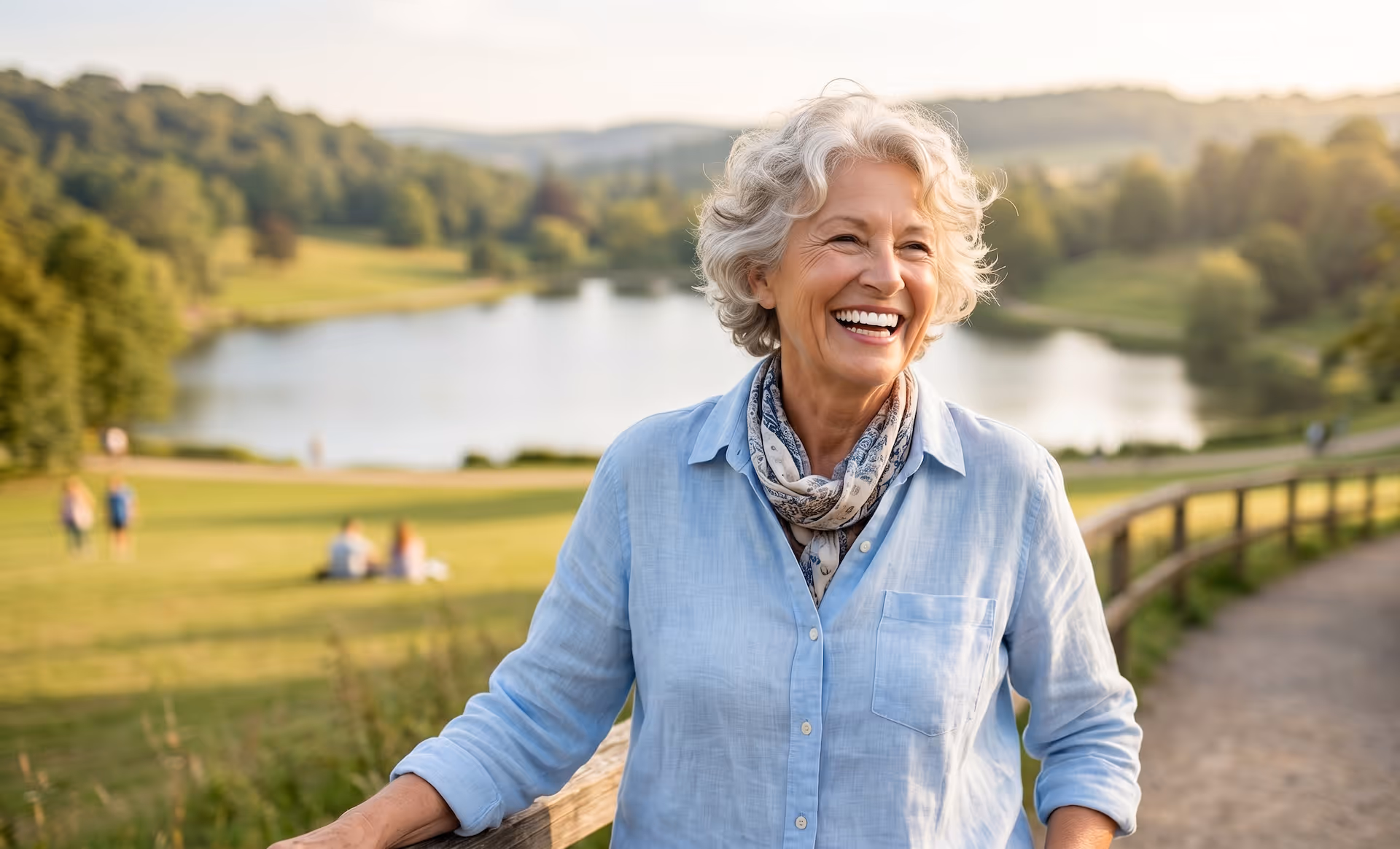 Smiling elderly woman with short gray hair standing outdoors by a wooden fence near a lake.