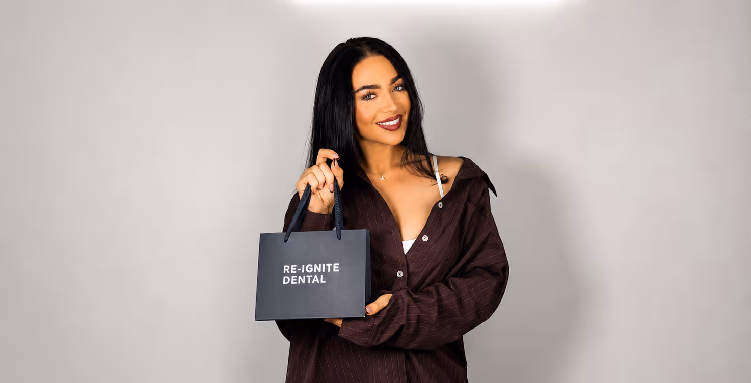 Smiling woman with long black hair holding a black Re-Ignite Dental shopping bag against a plain background.