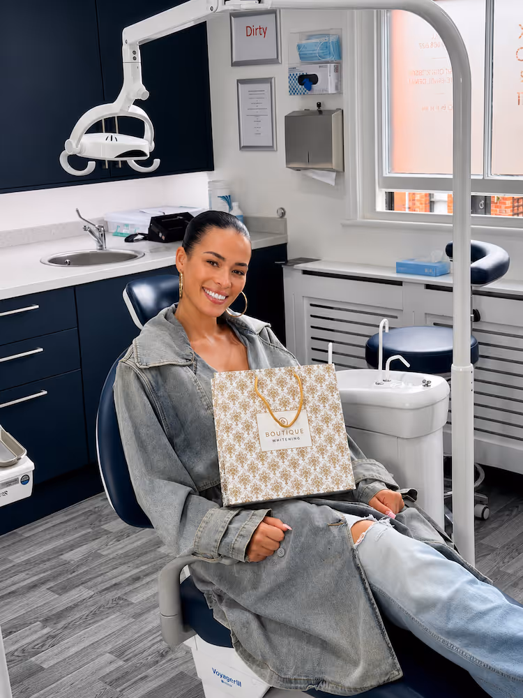 Smiling woman in a dental chair holding a Boutique Whitening paper bag in a bright dental office.