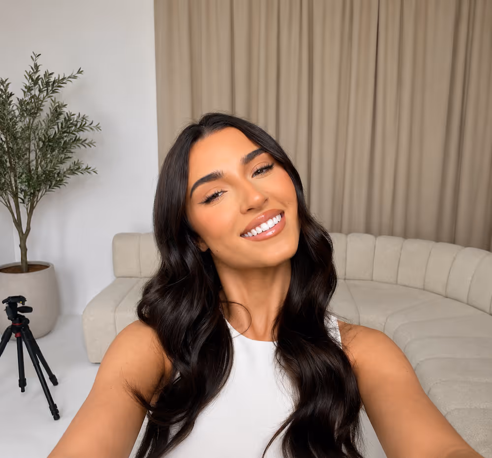 Young woman with long dark hair smiling and taking a selfie in a modern room with a beige curved sofa, a large plant, and a tripod.