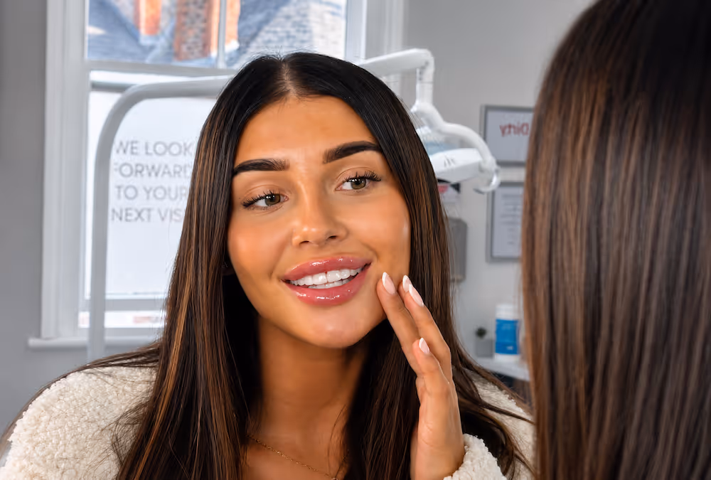Young woman with long dark hair smiling and touching her face in front of a mirror in a dental clinic.