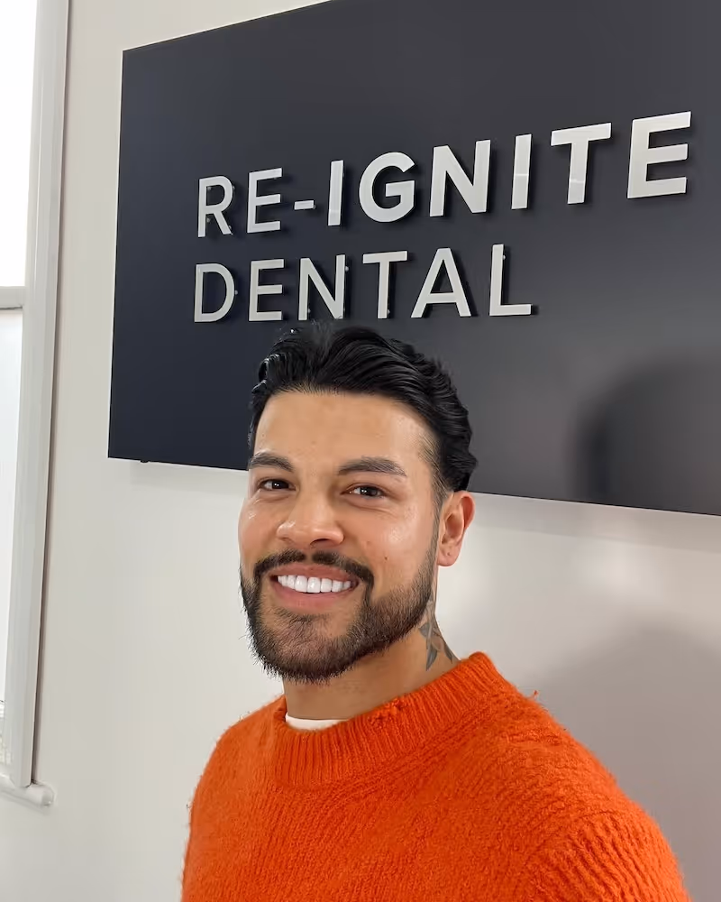 Smiling man in an orange sweater standing in front of a Re-Ignite Dental sign.