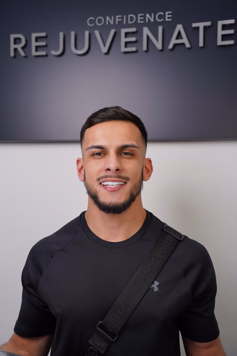 Smiling young man with short beard in black shirt and strap, standing before a sign saying Confidence Rejuvenate.