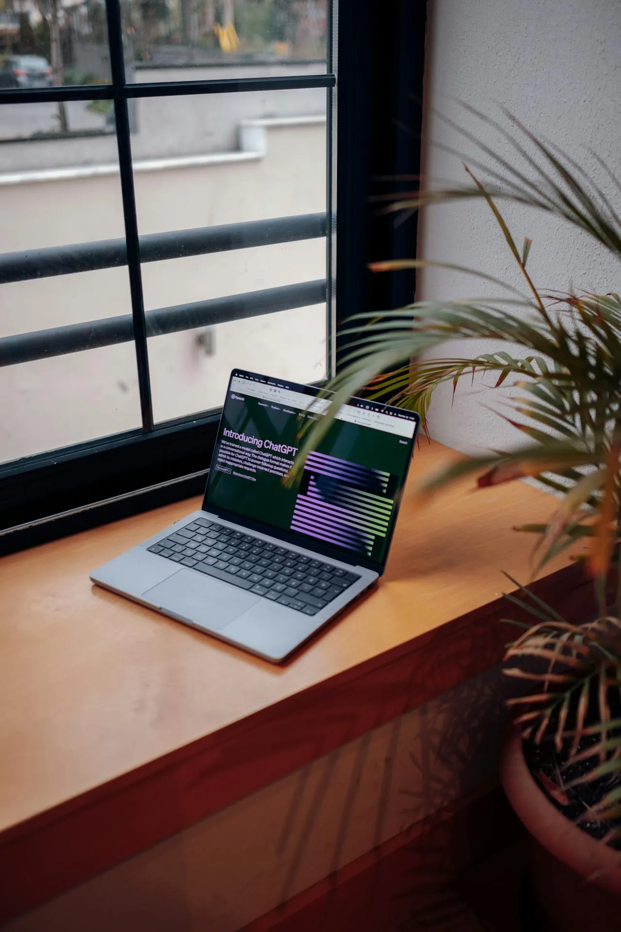Two men working on laptops at a shared desk with plants and computer monitors near a window.