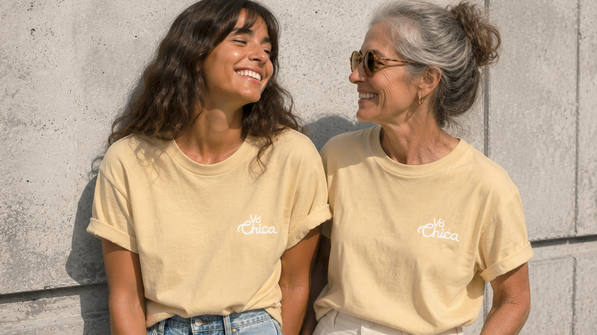 Two smiling women wearing matching pale yellow Vó Chica t-shirts sitting against a concrete wall.