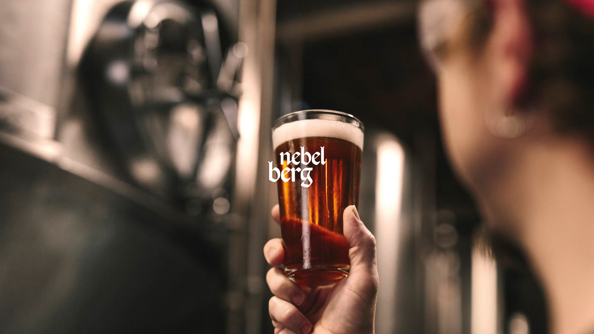 Hand holding a glass of amber beer with foam in a brewery setting.