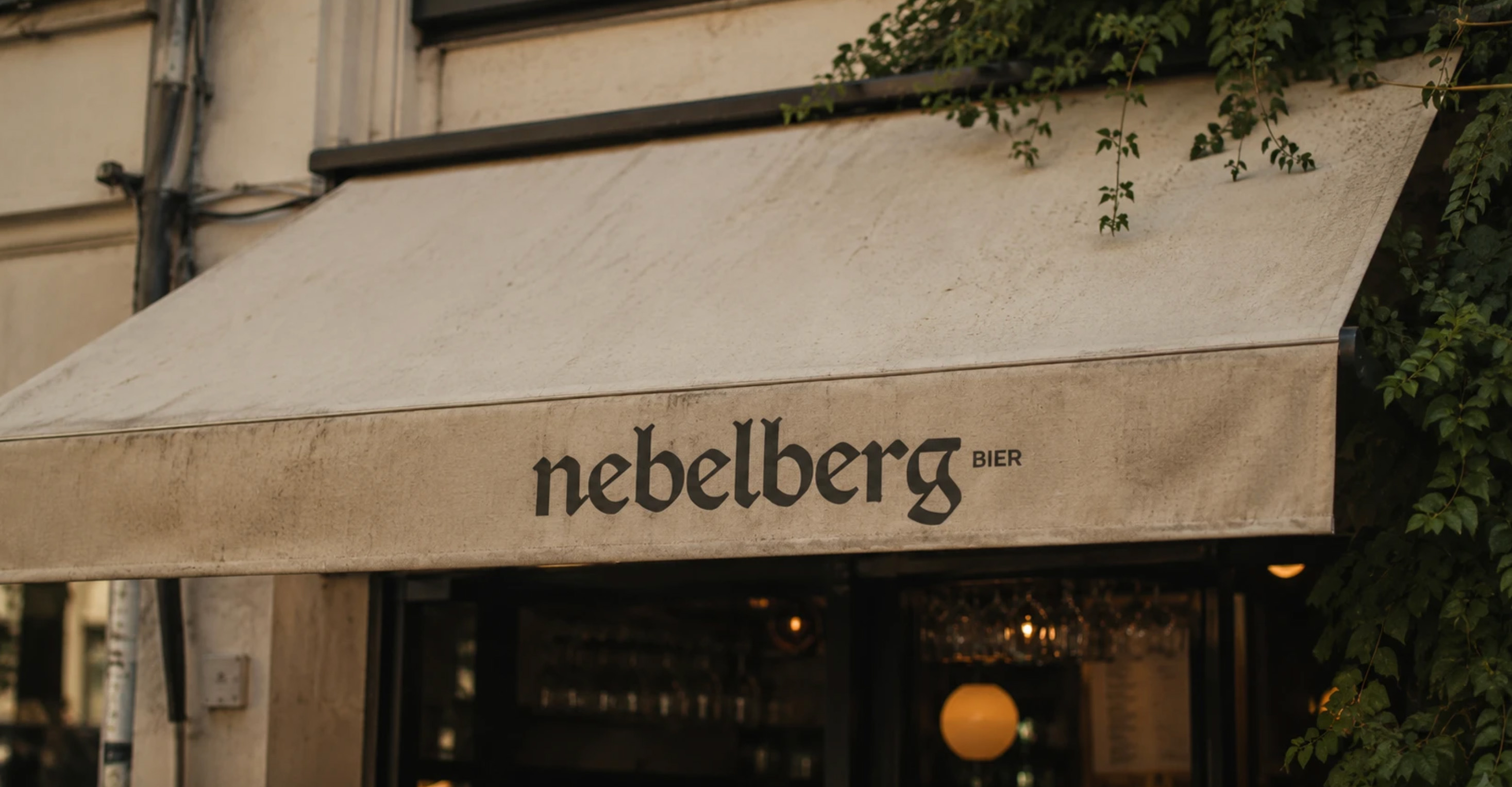 Outdoor beige awning with black text reading 'nebelberg BIER' above a bar entrance with hanging lights.