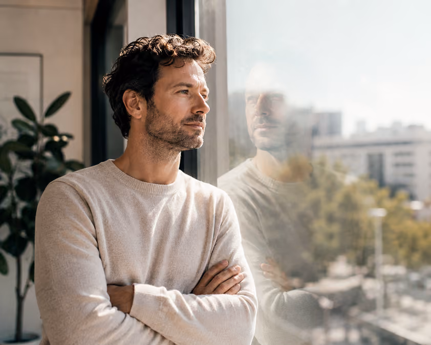 A man with curly hair and a beard is standing indoors with his arms crossed, looking thoughtfully out of a large window that reflects his image and shows an urban scene outside.