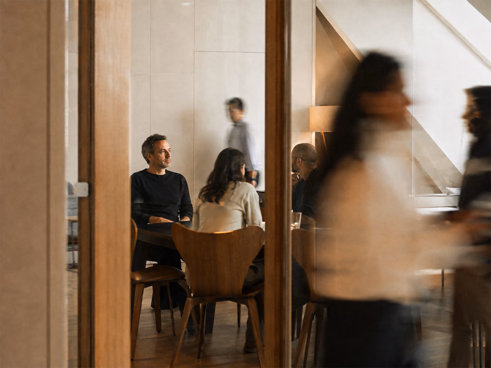 People sitting around a table in a modern indoor setting with blurred figures walking past in the foreground.