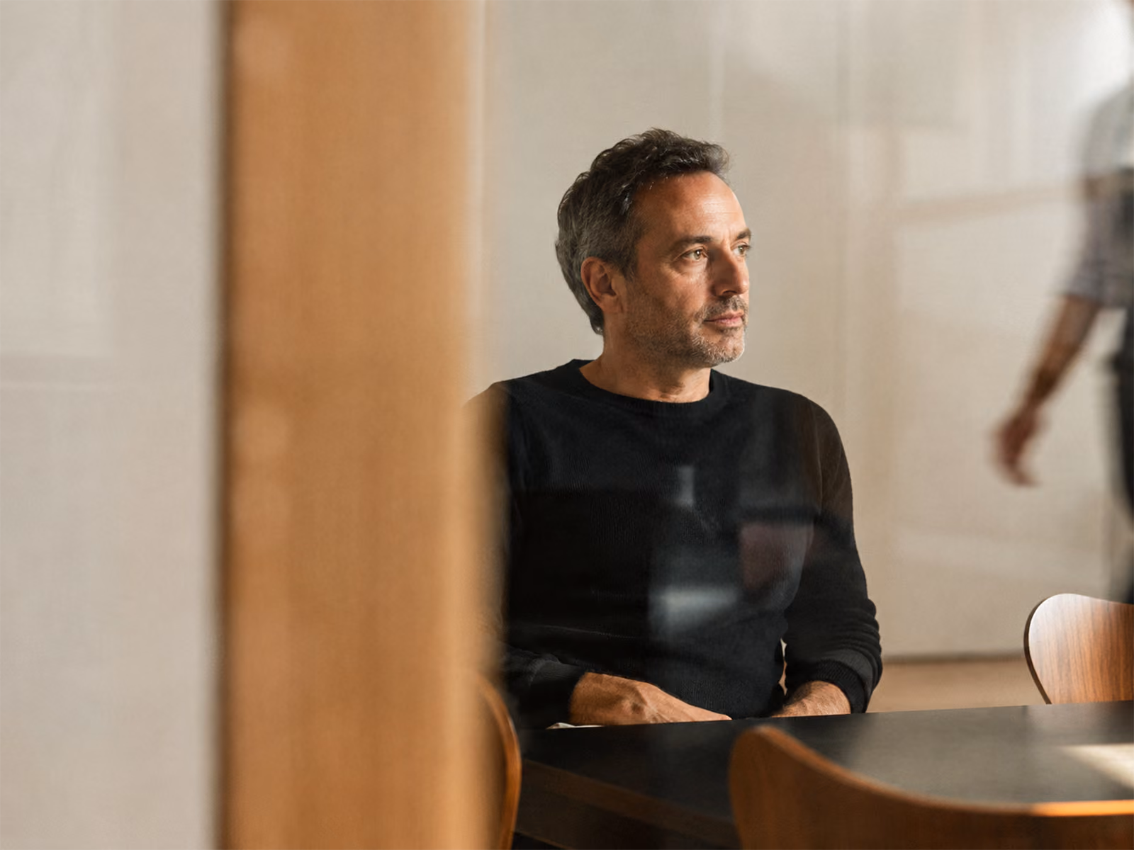 Middle-aged man with short dark hair and slight gray, wearing a black long-sleeve shirt, sitting at a table indoors, looking contemplatively to the right, with a blurred wooden panel and another person partially visible walking in the background.