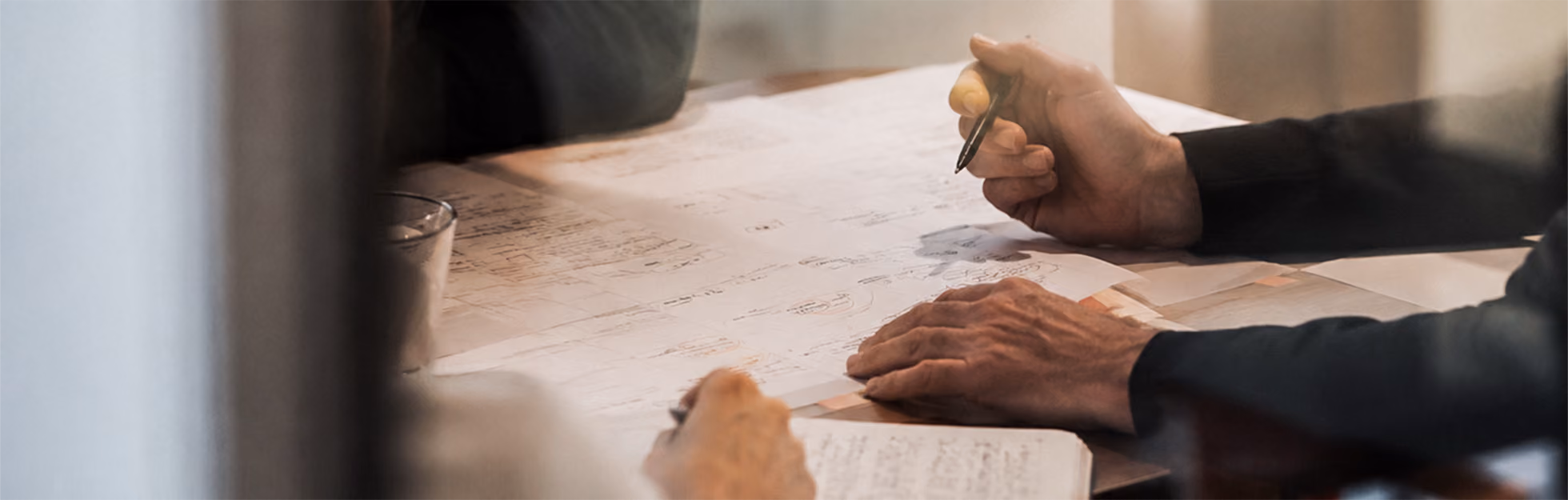 Two people at a table reviewing a large sheet of paper filled with handwritten notes, one person holding a pen over the paper, the other notes on a notepad.