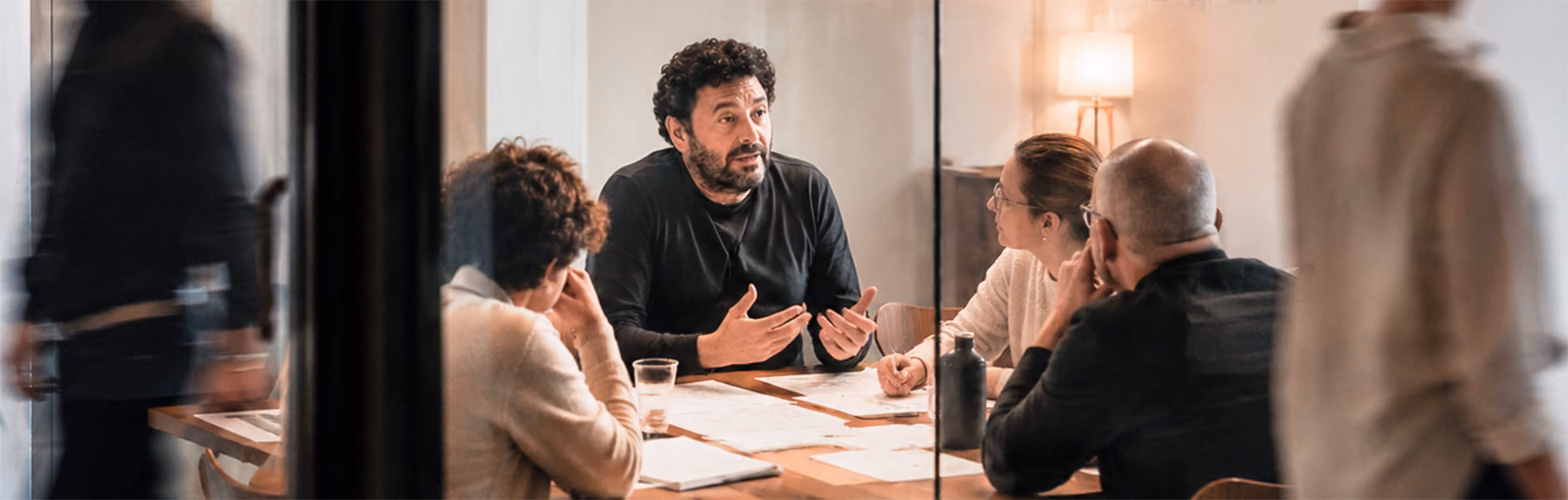 Four people seated around a table in a meeting room, with one man speaking and gesturing while others listen attentively.